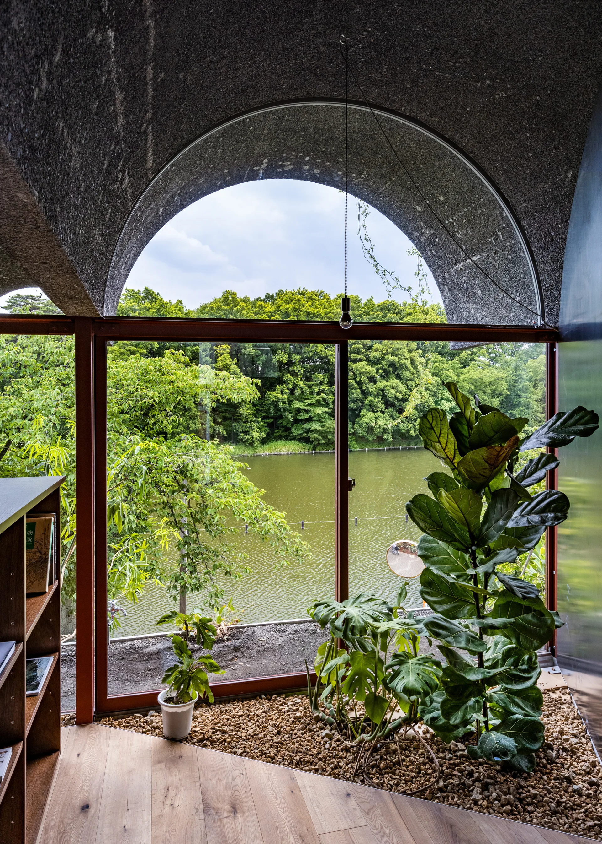 An interior space with a vaulted concrete ceiling features a large window overlooking a pond and an indoor gravel planter with houseplants.