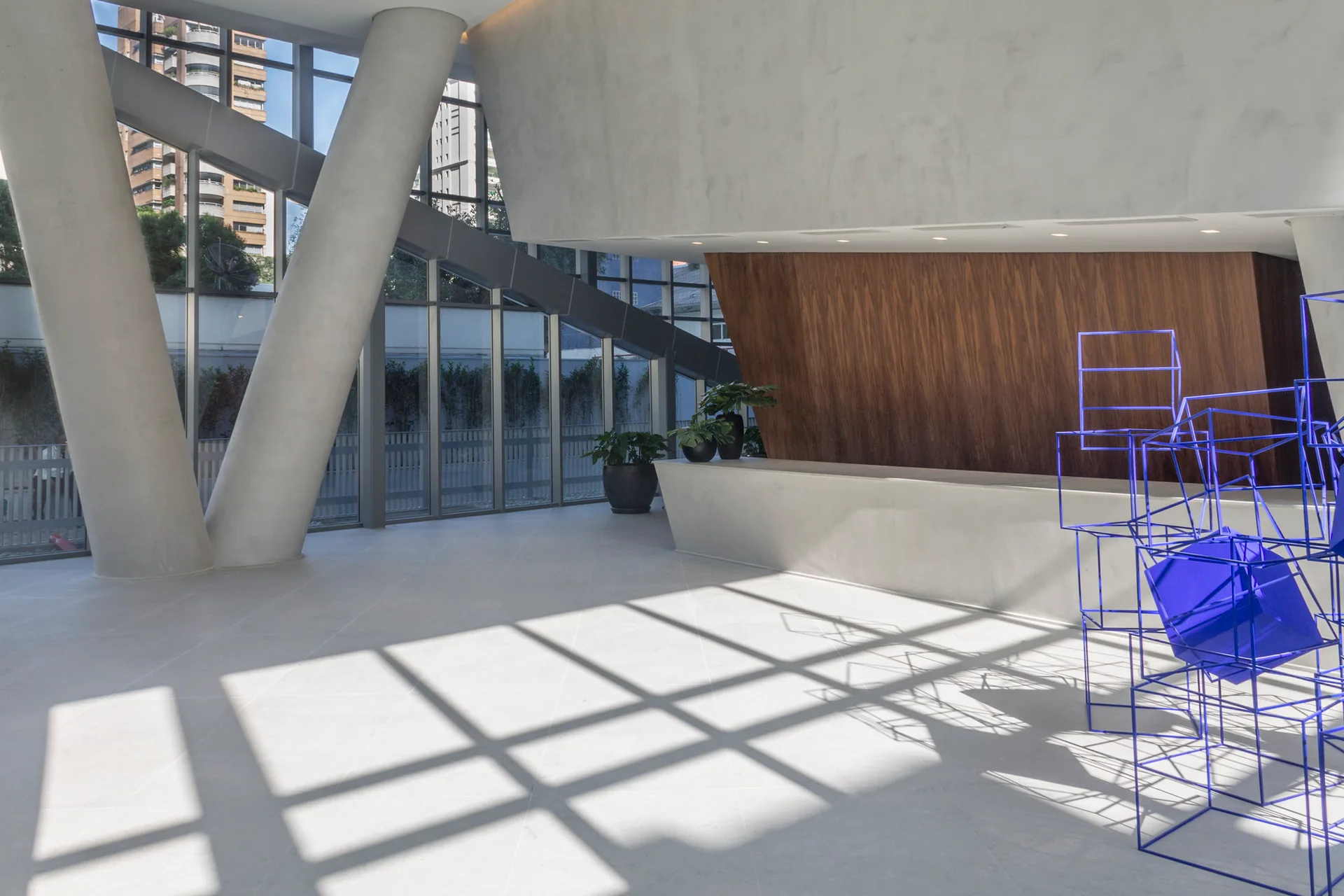 A spacious modern lobby features large angled concrete columns, floor-to-ceiling windows, and a white reception desk set against a wood-paneled wall.