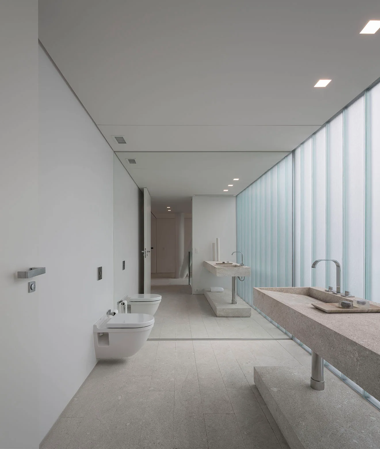 A minimalist bathroom featuring long textured stone vanities, wall-mounted toilets, and a large translucent channel glass wall.