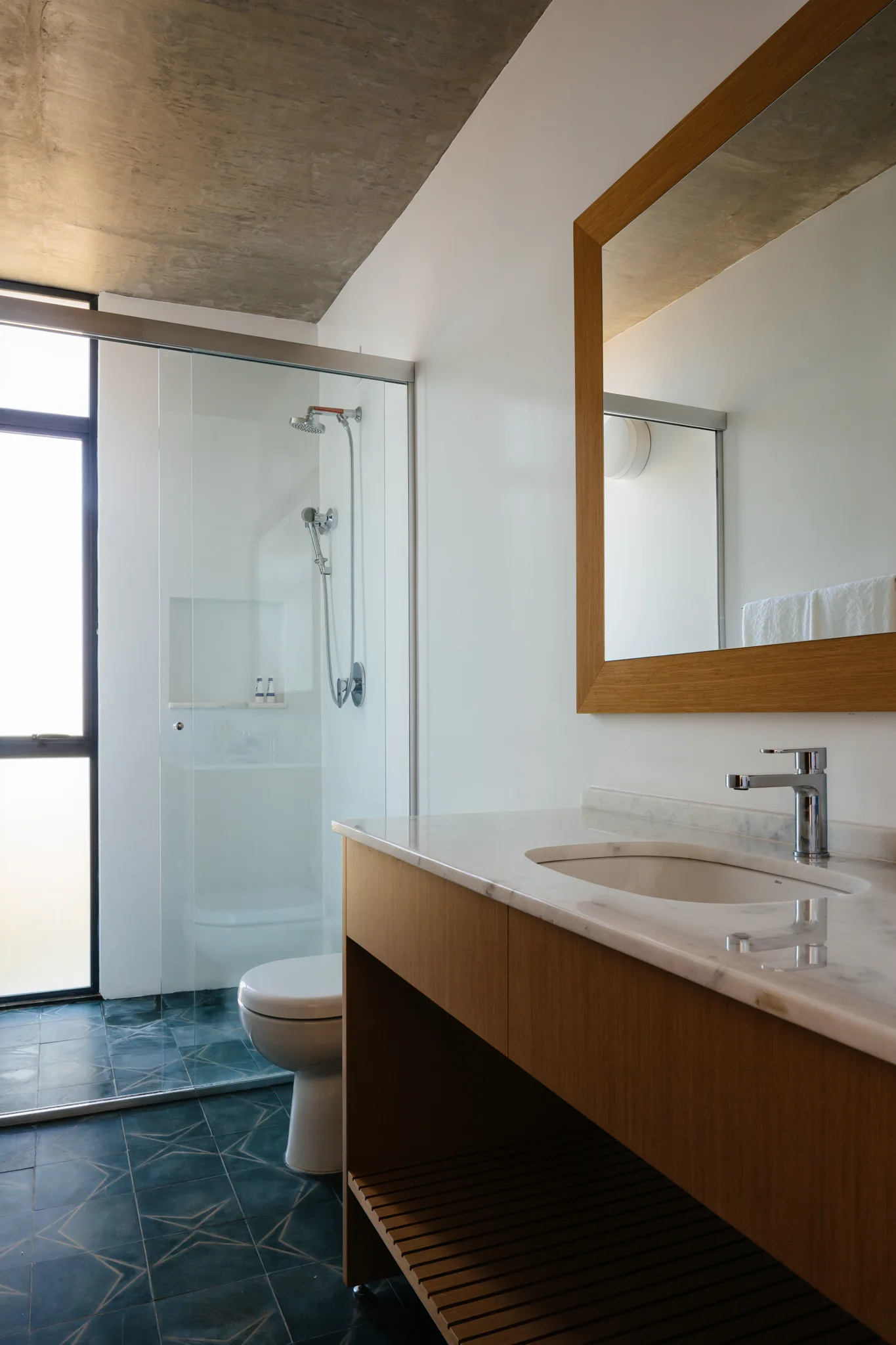 A modern bathroom features a wood vanity with a marble countertop, a wood-framed mirror, a glass shower stall, and an exposed concrete ceiling.