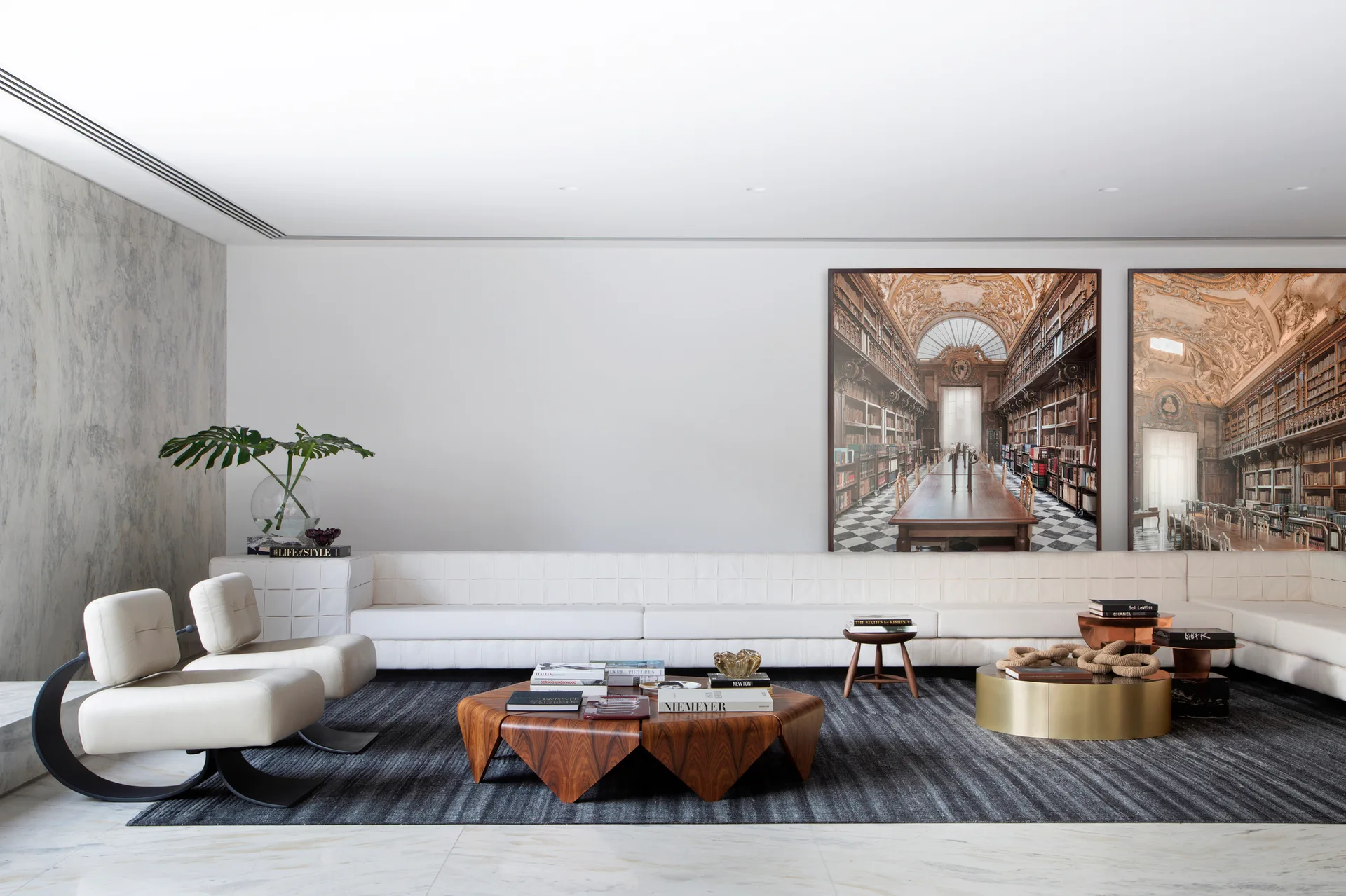 A minimalist living room with a long white modular sofa, a faceted wood coffee table, and large-scale architectural prints on a white wall.