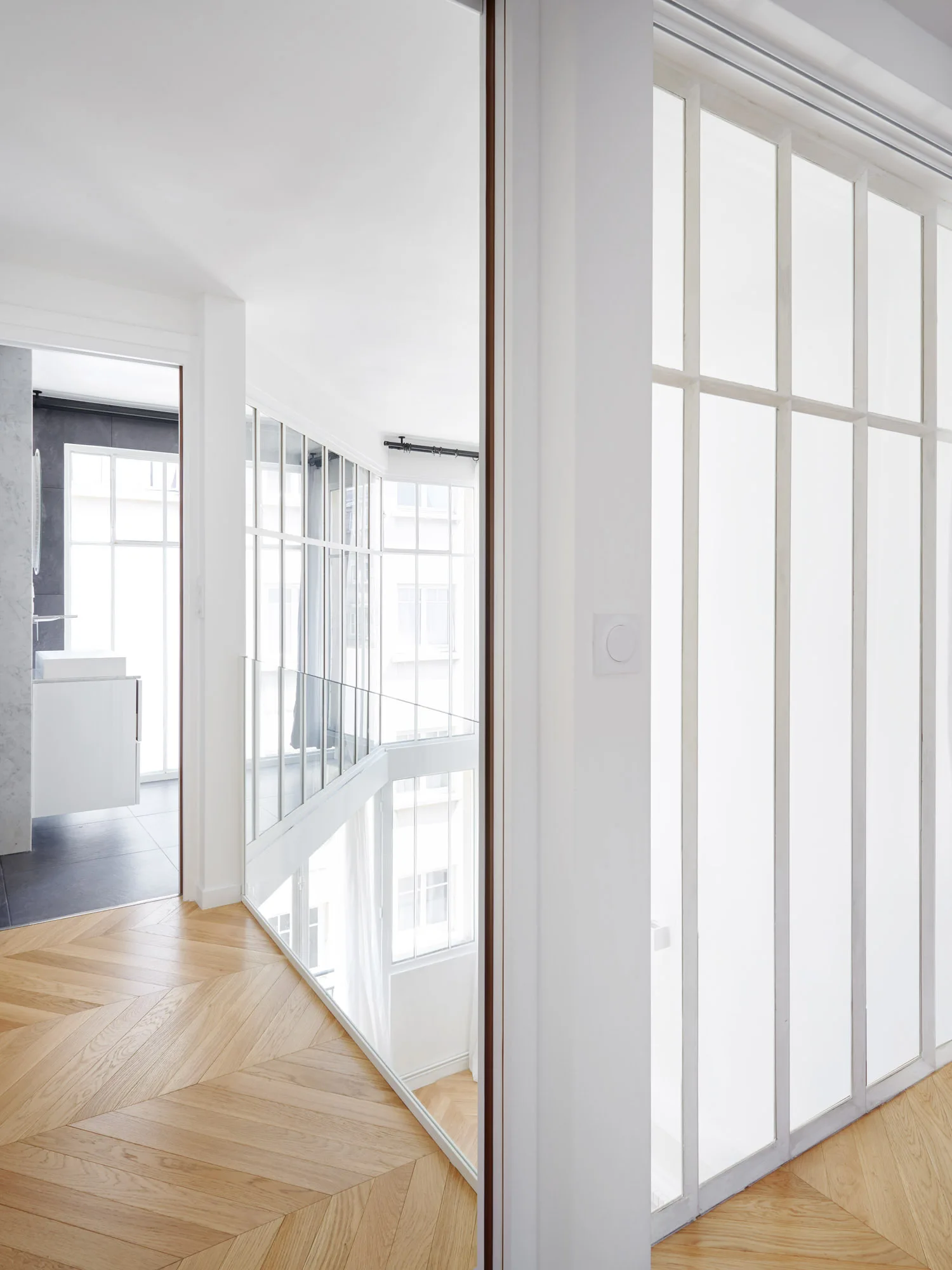 A bright hallway featuring light wood chevron flooring, white walls, and a large white-framed glass partition overlooking a lower level.