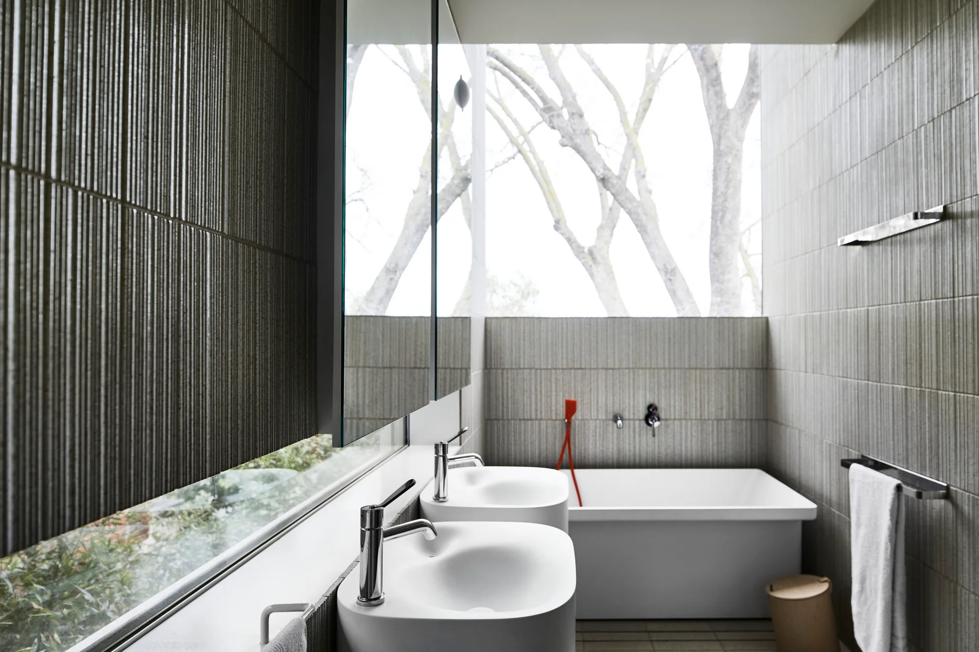 Minimalist bathroom featuring grey ribbed tile walls, a white double vanity, a rectangular bathtub, and a window displaying tree branches.