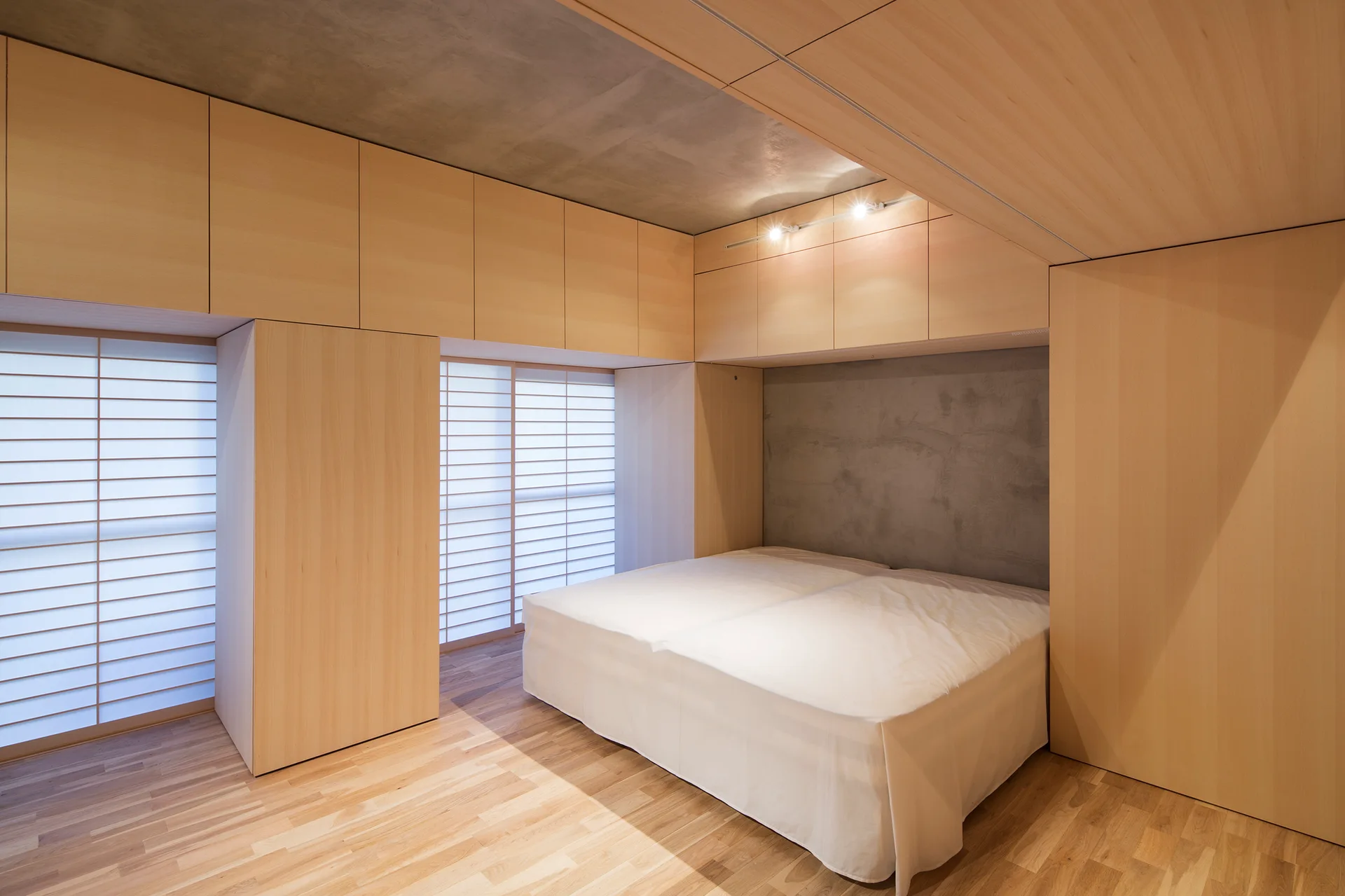 A minimalist bedroom features light wood built-in wall cabinets, a bed with white sheets, a raw concrete ceiling, and sliding shoji-style screens.