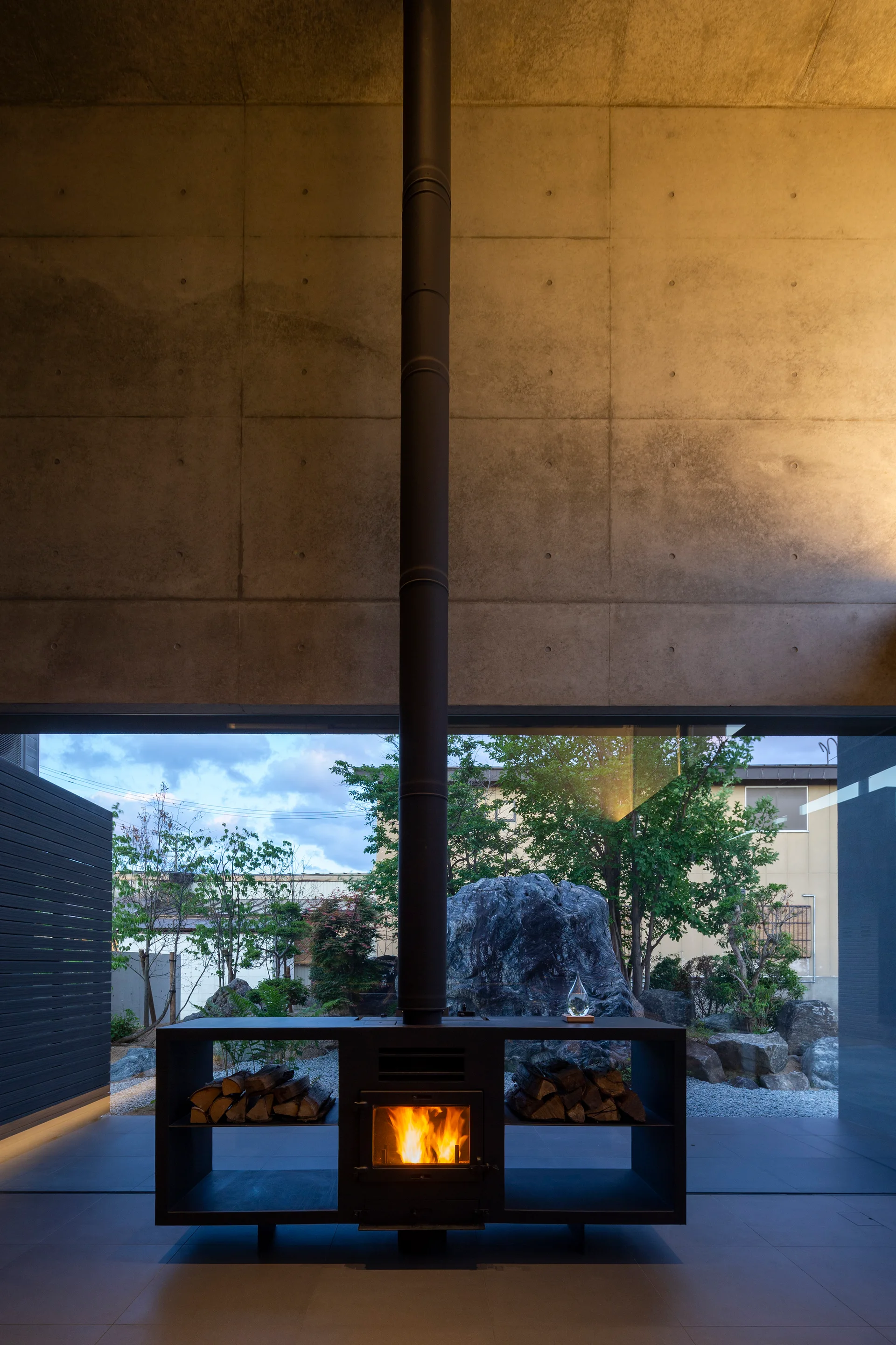 A black metal fireplace with integrated wood storage stands before a glass window overlooking a garden beneath a tall raw concrete wall.