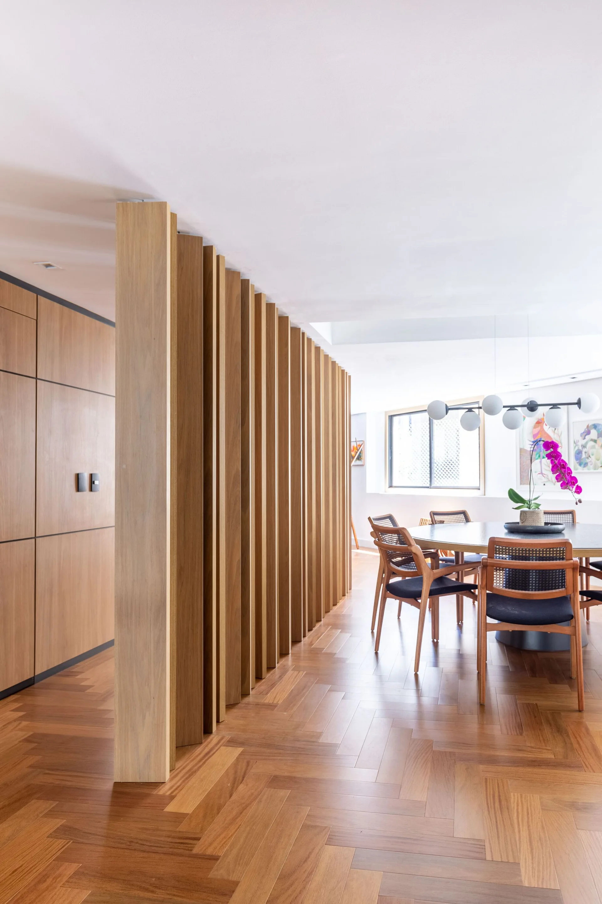 A modern dining room features a round table with cane chairs next to a vertical wood slat partition and herringbone wood flooring.