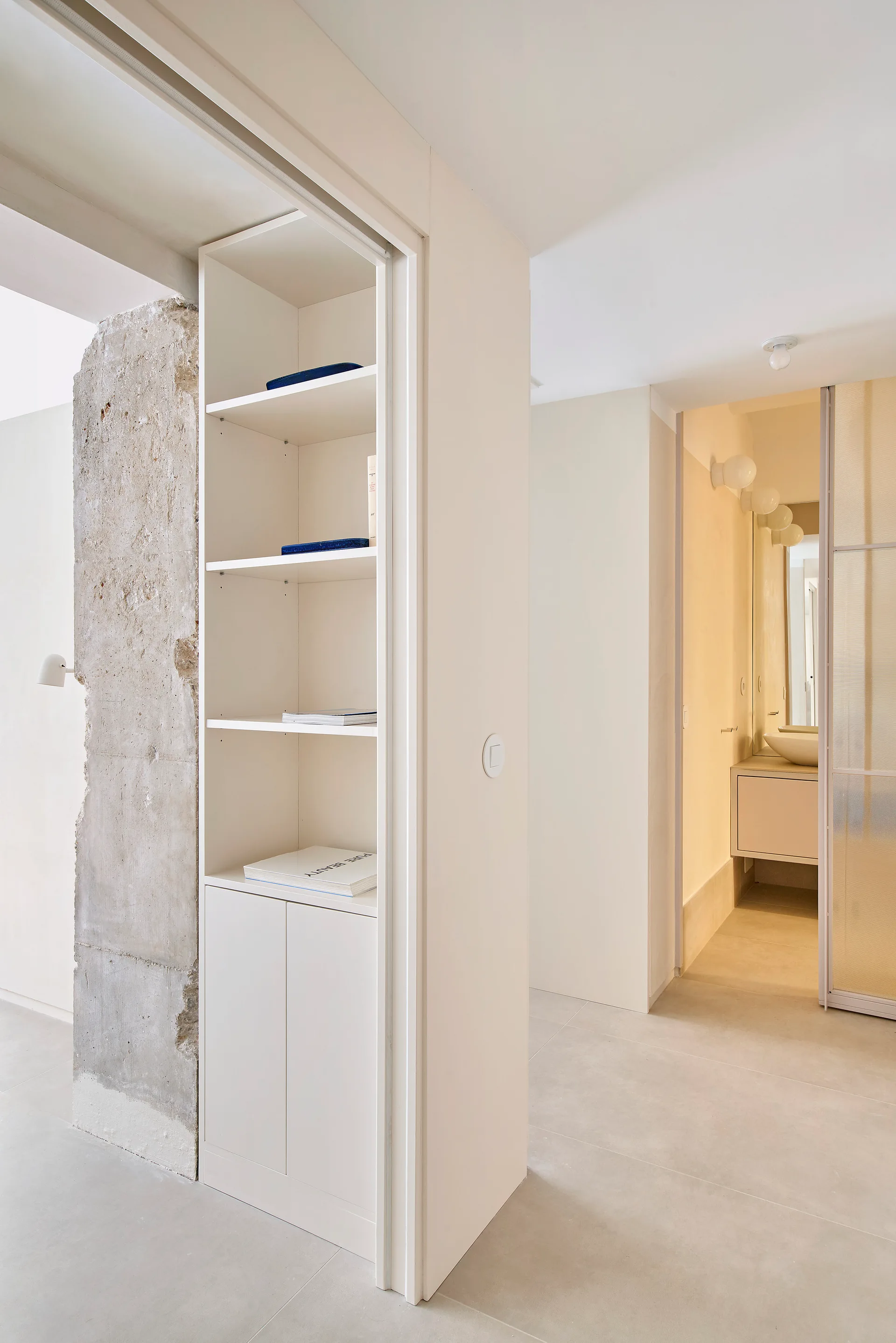 A tall white built-in bookcase stands next to an exposed concrete pillar, with a view into a minimalist bathroom in the background.