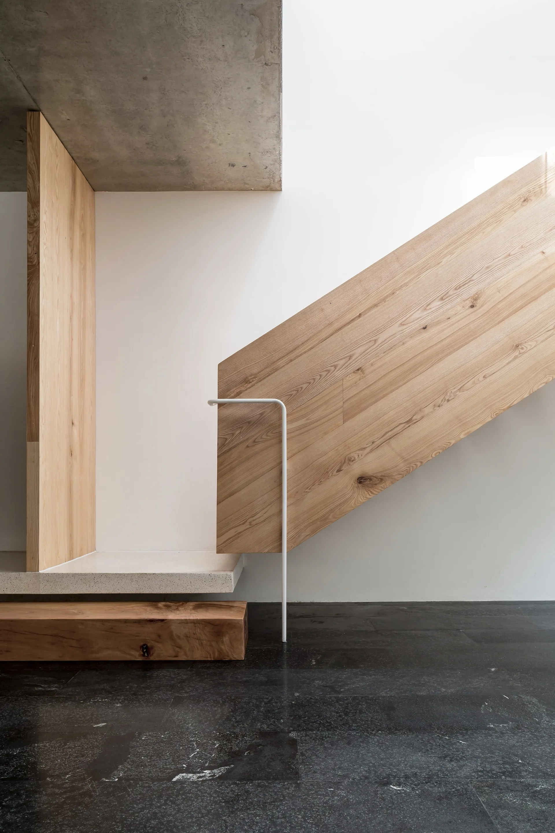 A minimalist staircase features light wood paneling, a thin white metal handrail, a raw concrete ceiling, and a dark stone tiled floor.