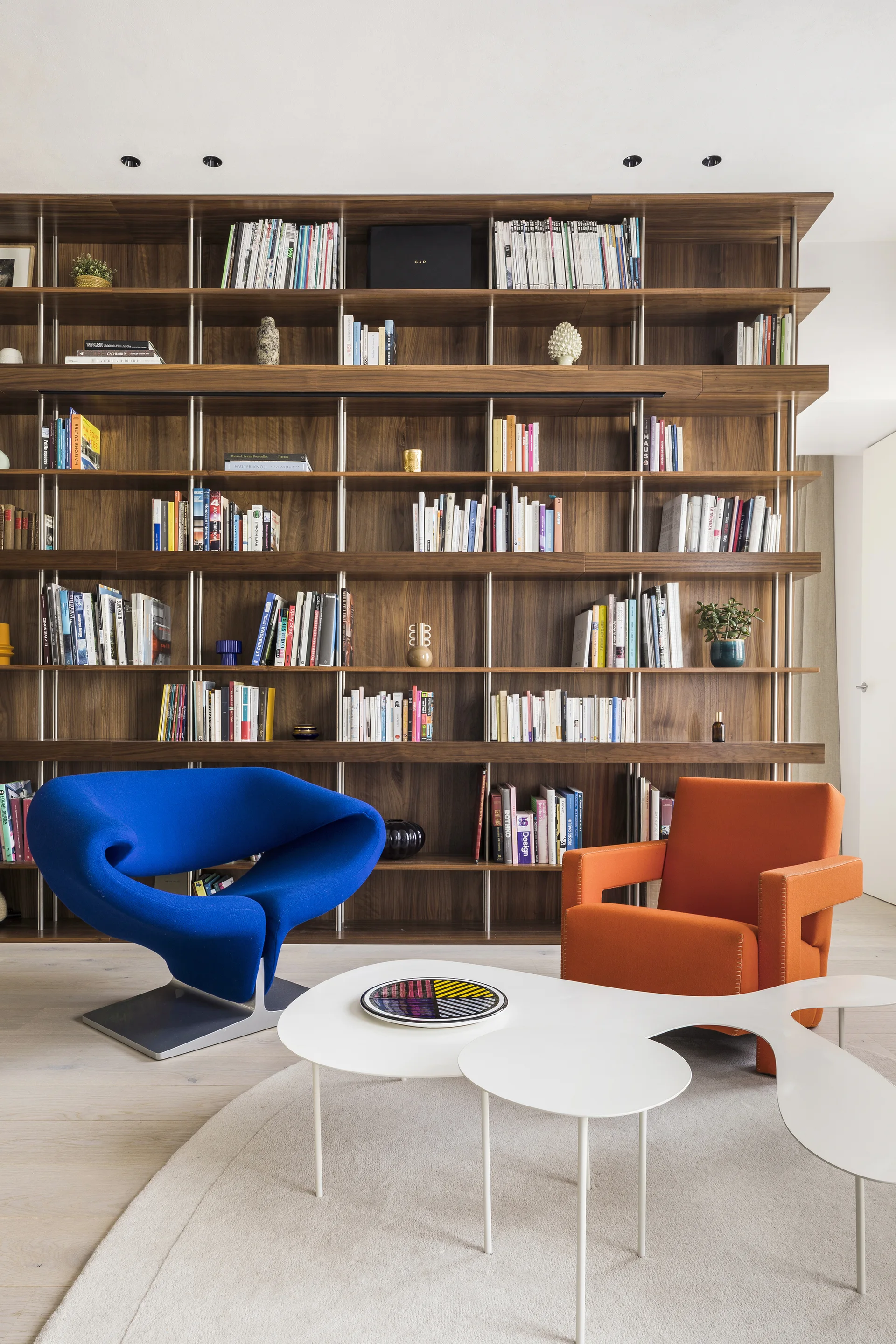 A large wall-to-wall wooden bookshelf filled with books sits behind a sculptural blue armchair, an orange armchair, and a tiered white coffee table.