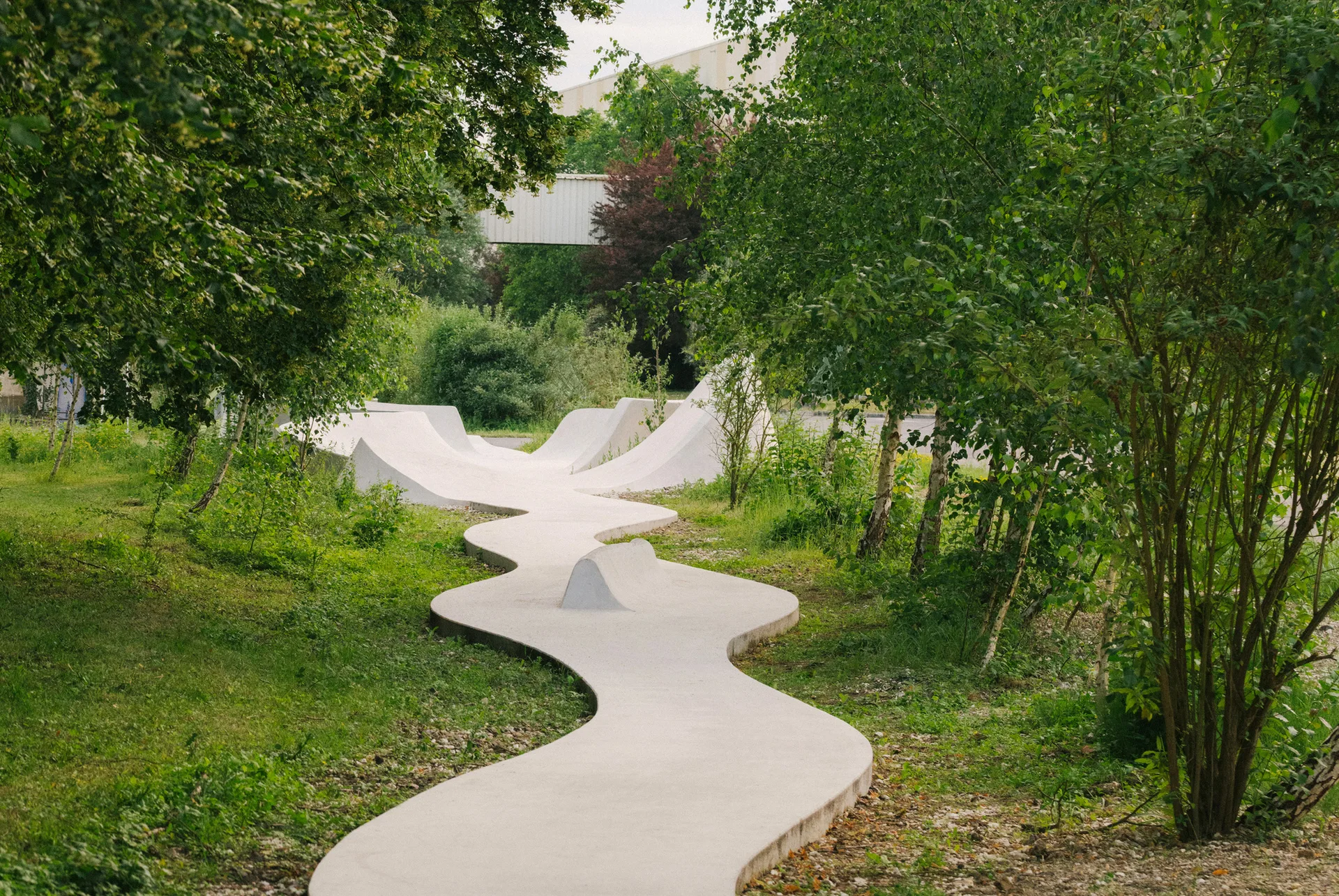 A winding white concrete pathway and sculptural ramp curves through a lush green park surrounded by tall trees.