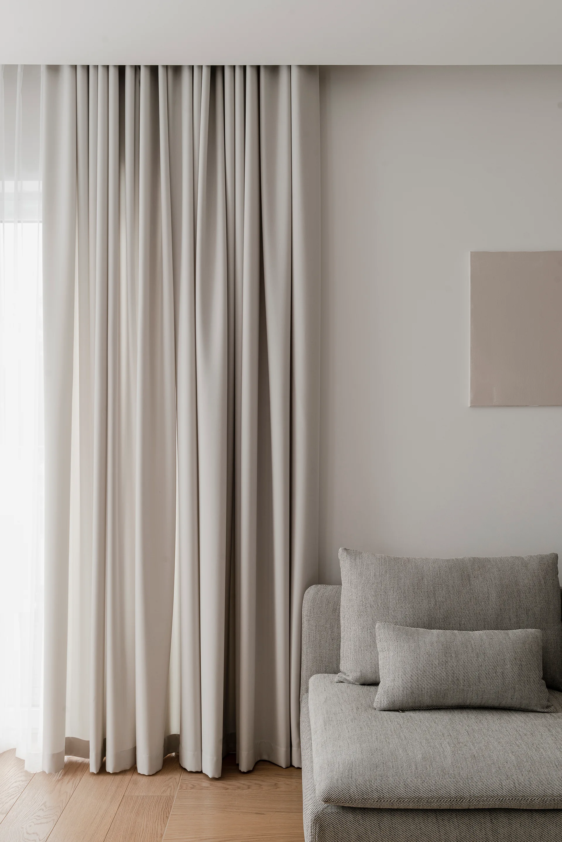 A textured gray sofa with matching pillows sits next to floor-to-ceiling beige curtains in a minimalist room with light wood floors.