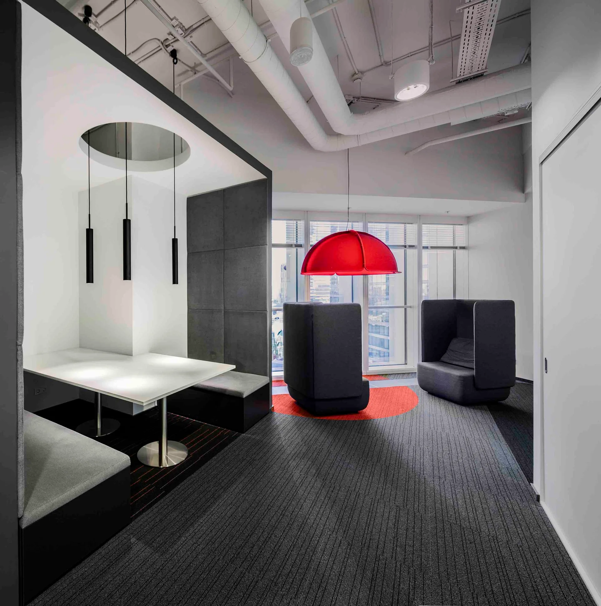A modern office break-out area features gray built-in booth seating, tall privacy chairs, and a red dome pendant light.