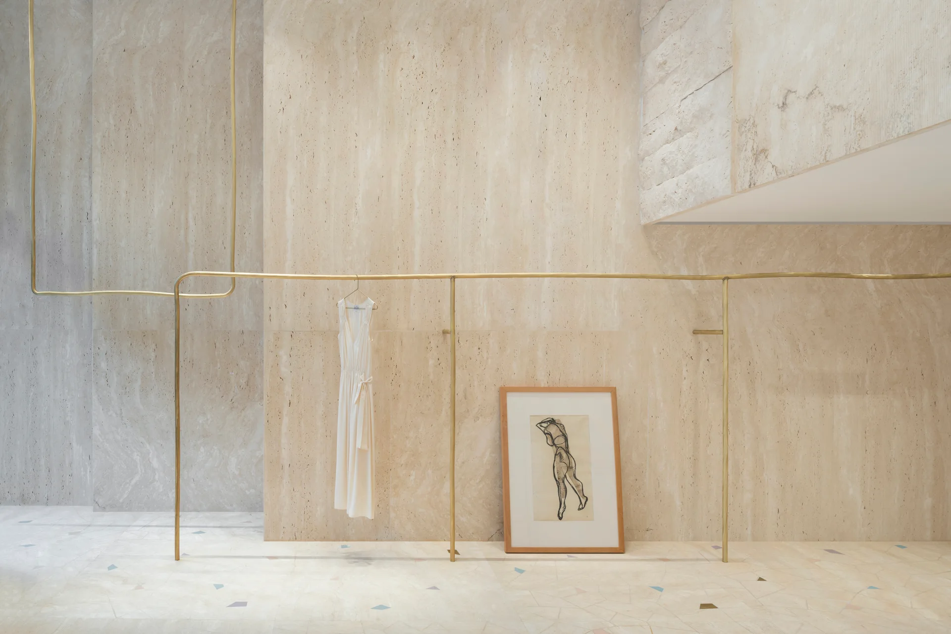 A thin brass garment rail runs along a beige travertine stone wall, with a single white dress hanging and a framed art piece leaning on the floor.