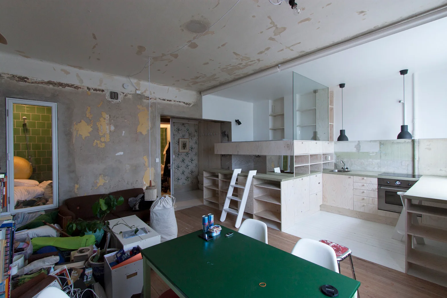 An open plan kitchen featuring custom plywood cabinetry, a matching loft bed with a white ladder, a green dining table, and unfinished concrete walls.