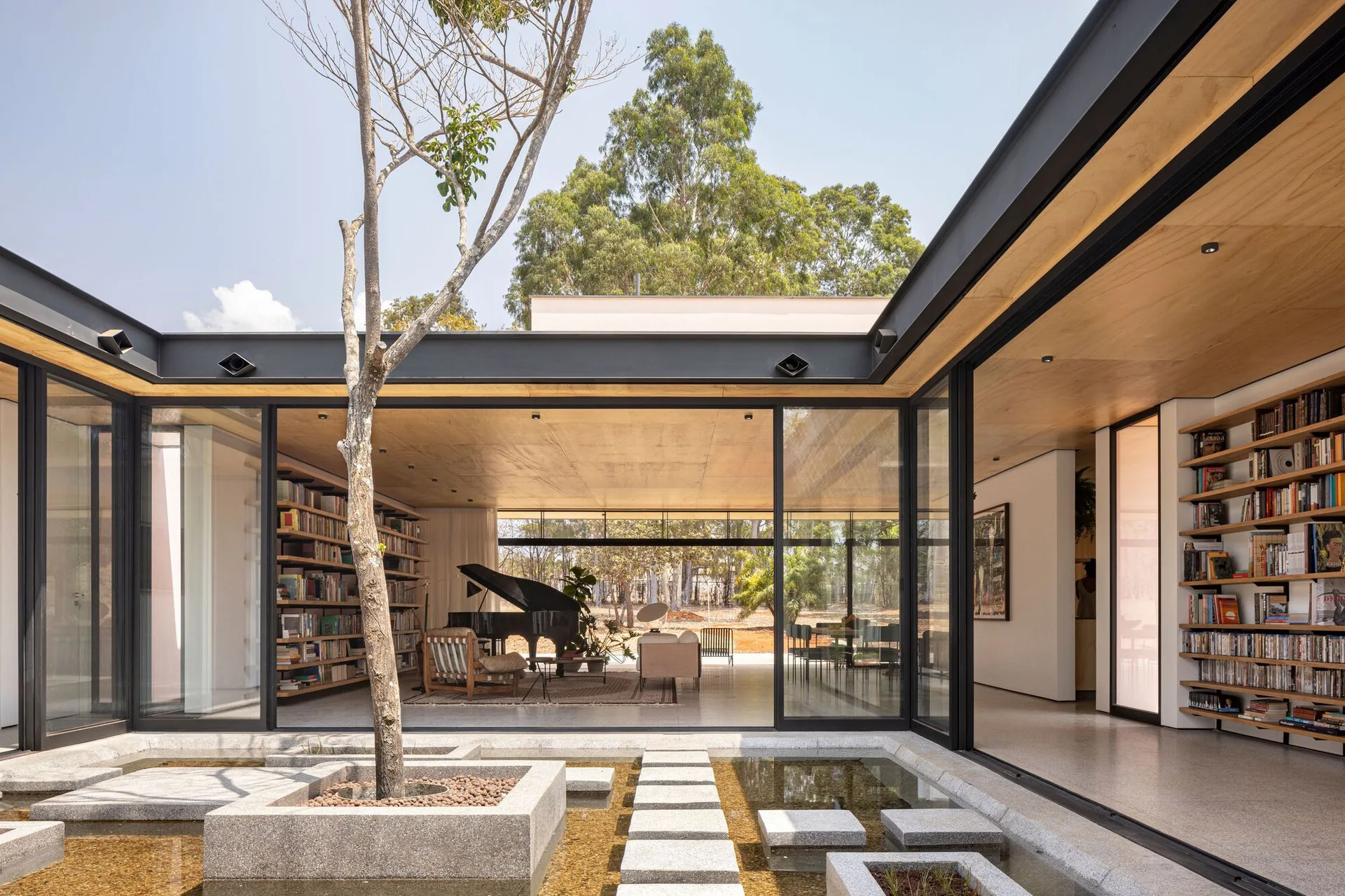 A modern residential courtyard features a reflecting pool with concrete stepping stones next to an open living area with floor-to-ceiling glass doors.