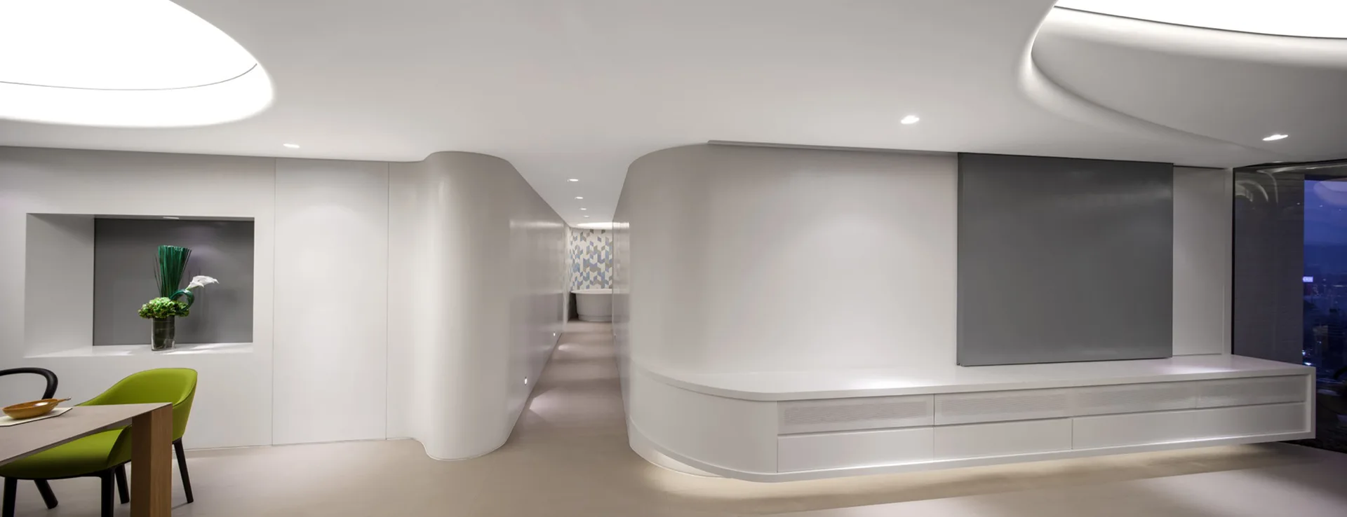 A minimalist white interior features organic curved walls, built-in storage console, recessed cove lighting, and a lime green dining chair.