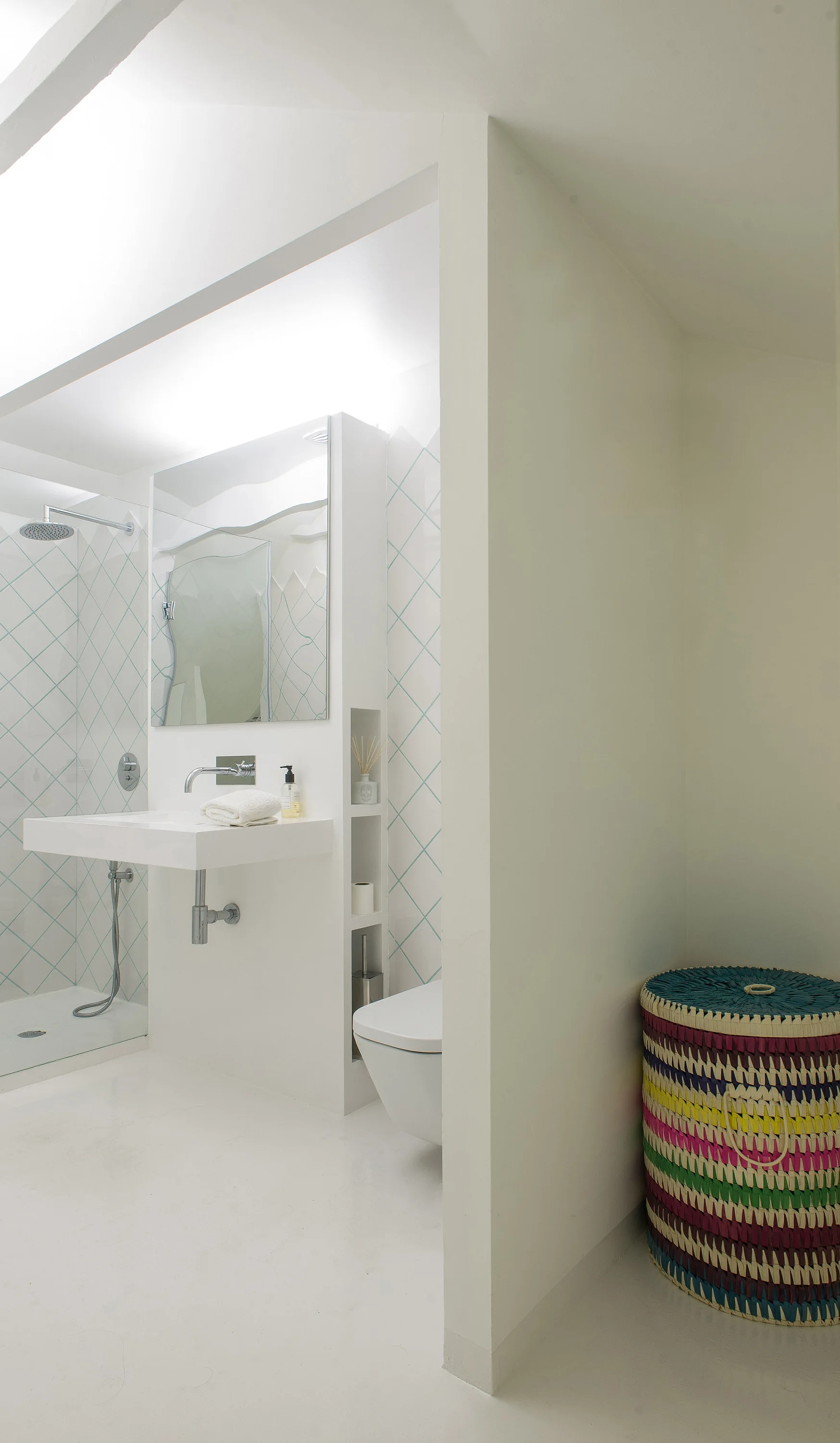 A minimalist white bathroom features a floating sink, a glass-enclosed shower, wall tiles with teal grout, and a colorful woven basket.