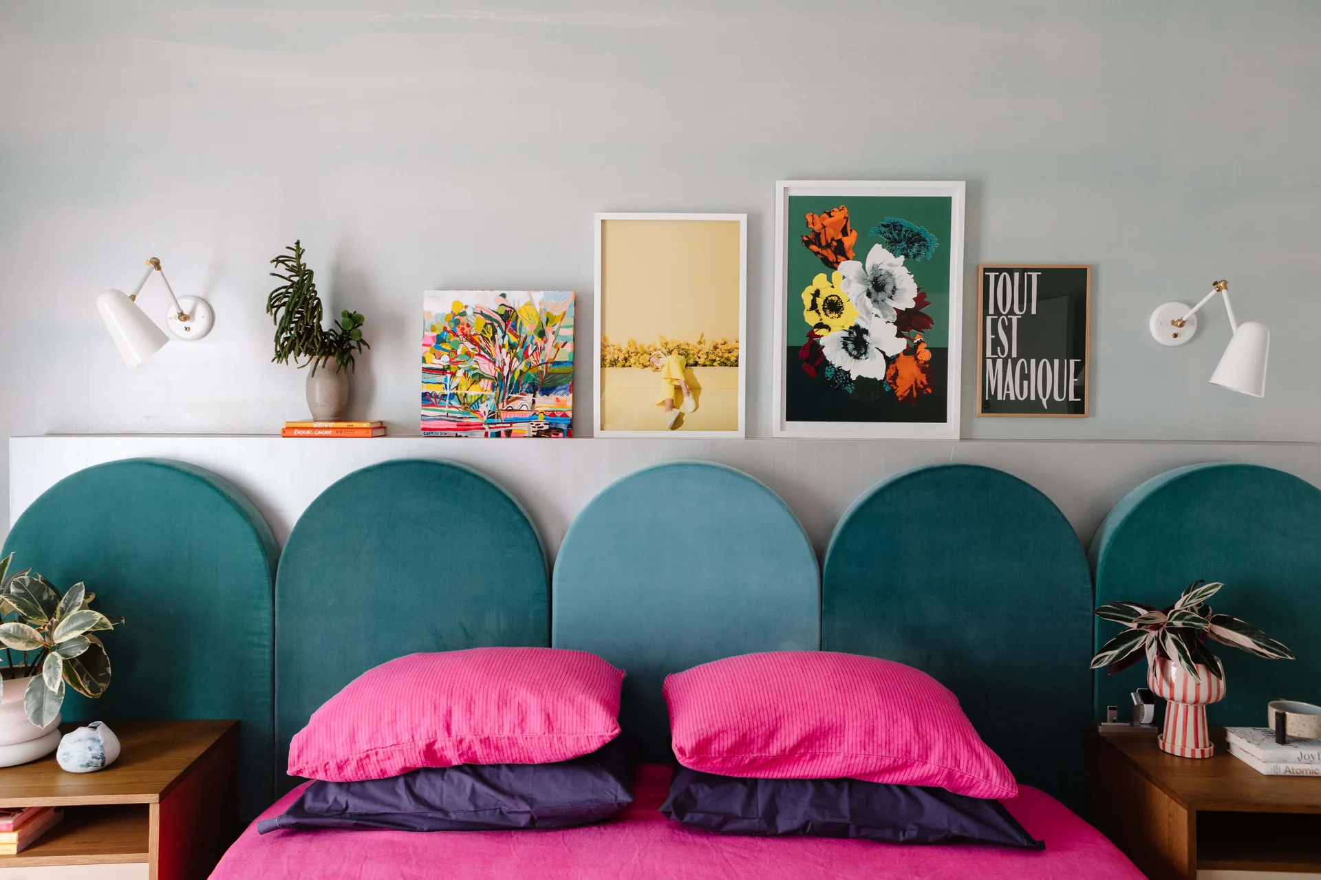 A teal velvet arched headboard sits behind pink and purple pillows, with a picture ledge above displaying framed artwork and white wall sconces.