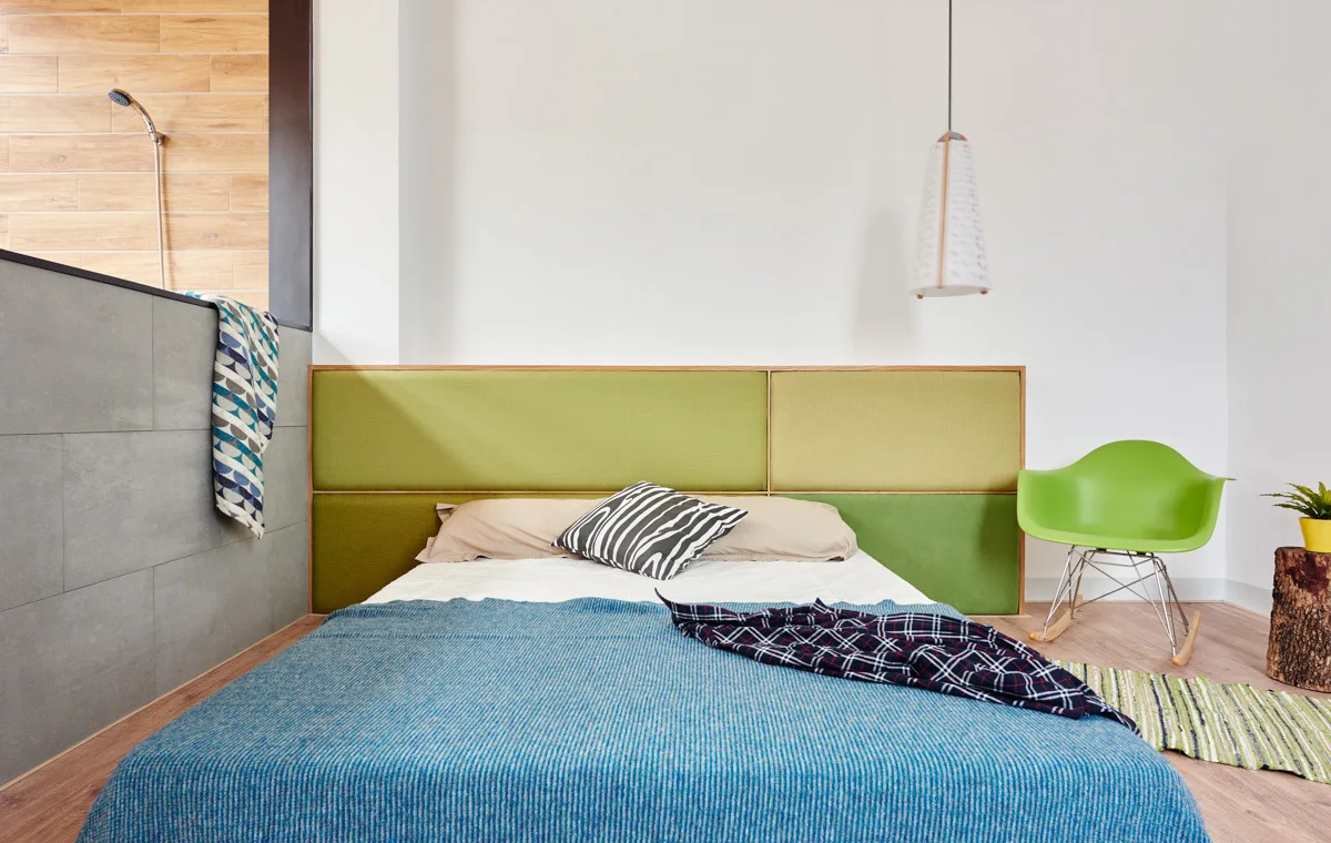 A modern bedroom features a bed with a green upholstered headboard, a blue bedspread, and a low grey tiled wall partitioning an open shower area.