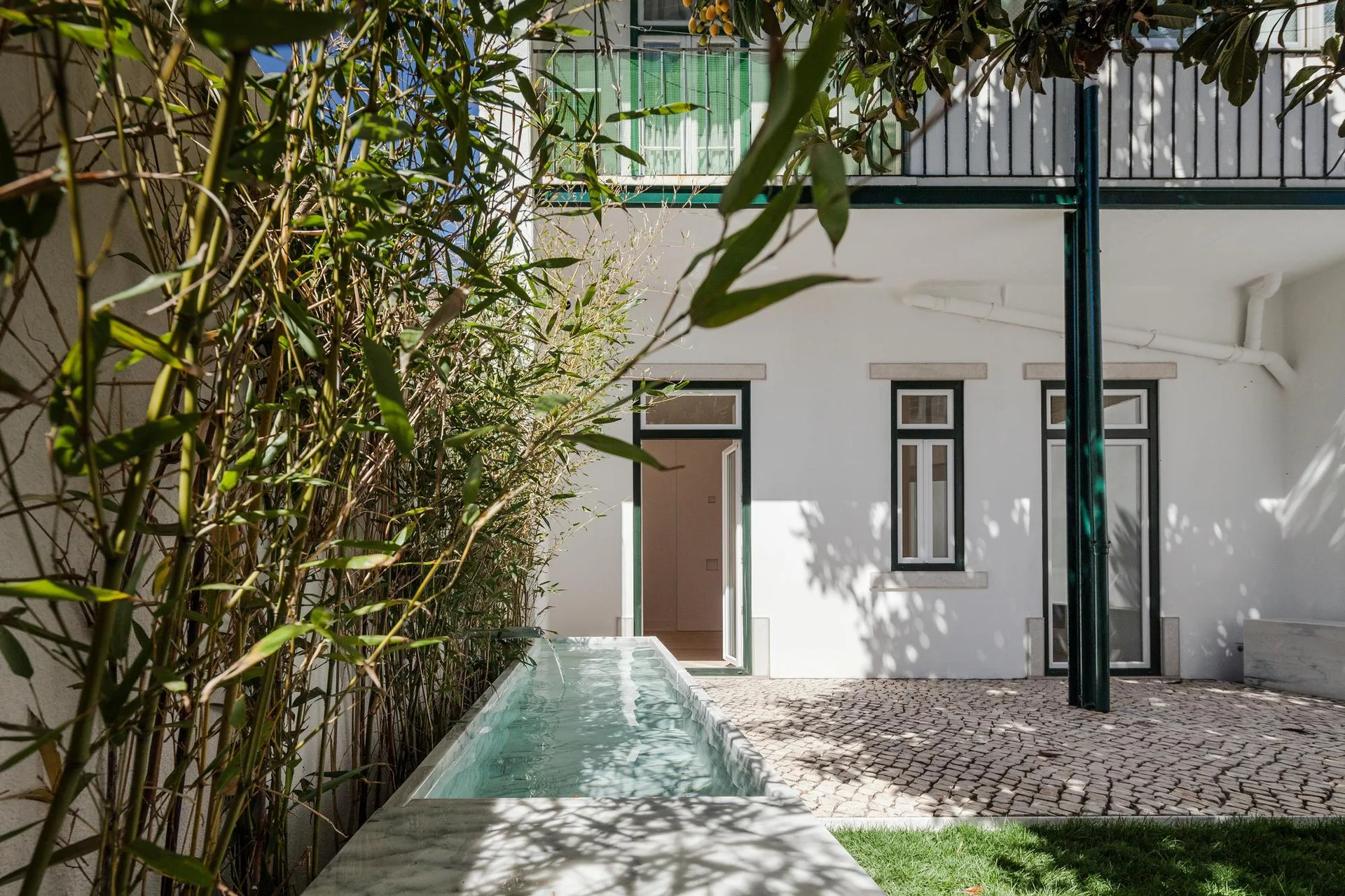 An outdoor courtyard features a narrow stone water basin beside tall bamboo plants, cobblestone flooring, and a white building facade.