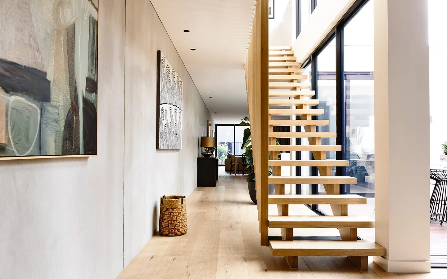 A modern hallway features a floating wood staircase with vertical slat railings adjacent to large floor-to-ceiling windows.