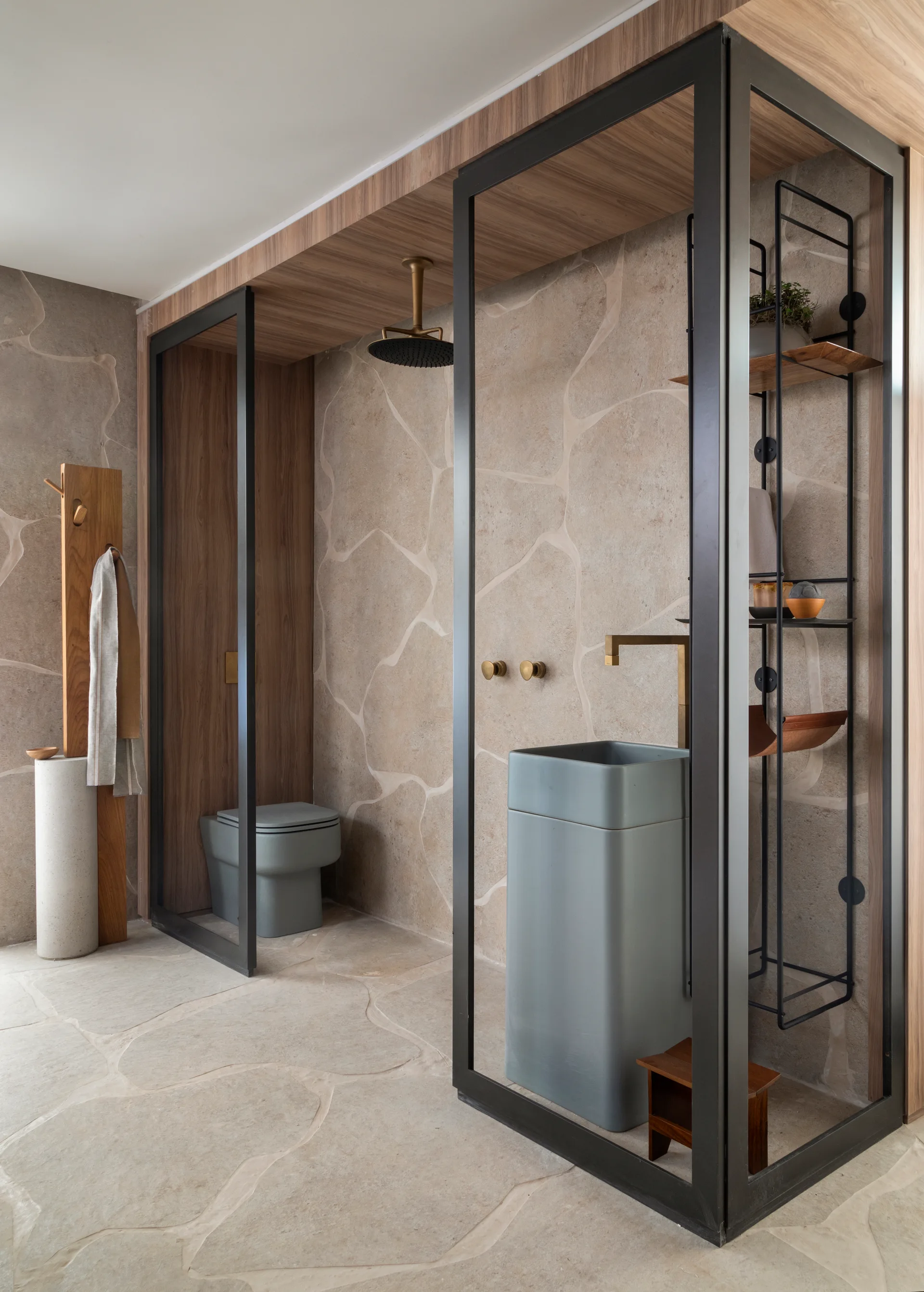 A minimalist bathroom features irregular flagstone wall panels, a dark wood ceiling, a grey pedestal sink, and a black metal partition frame.