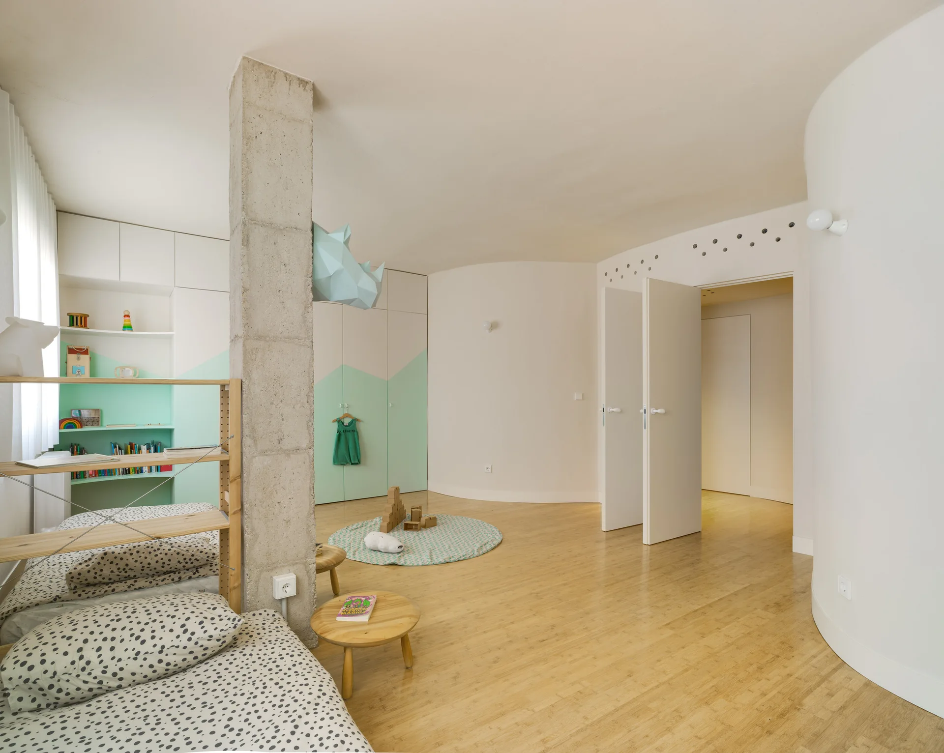 A children's bedroom features an exposed concrete column, curved white walls, light wood flooring, and built-in white and mint-green cabinets.