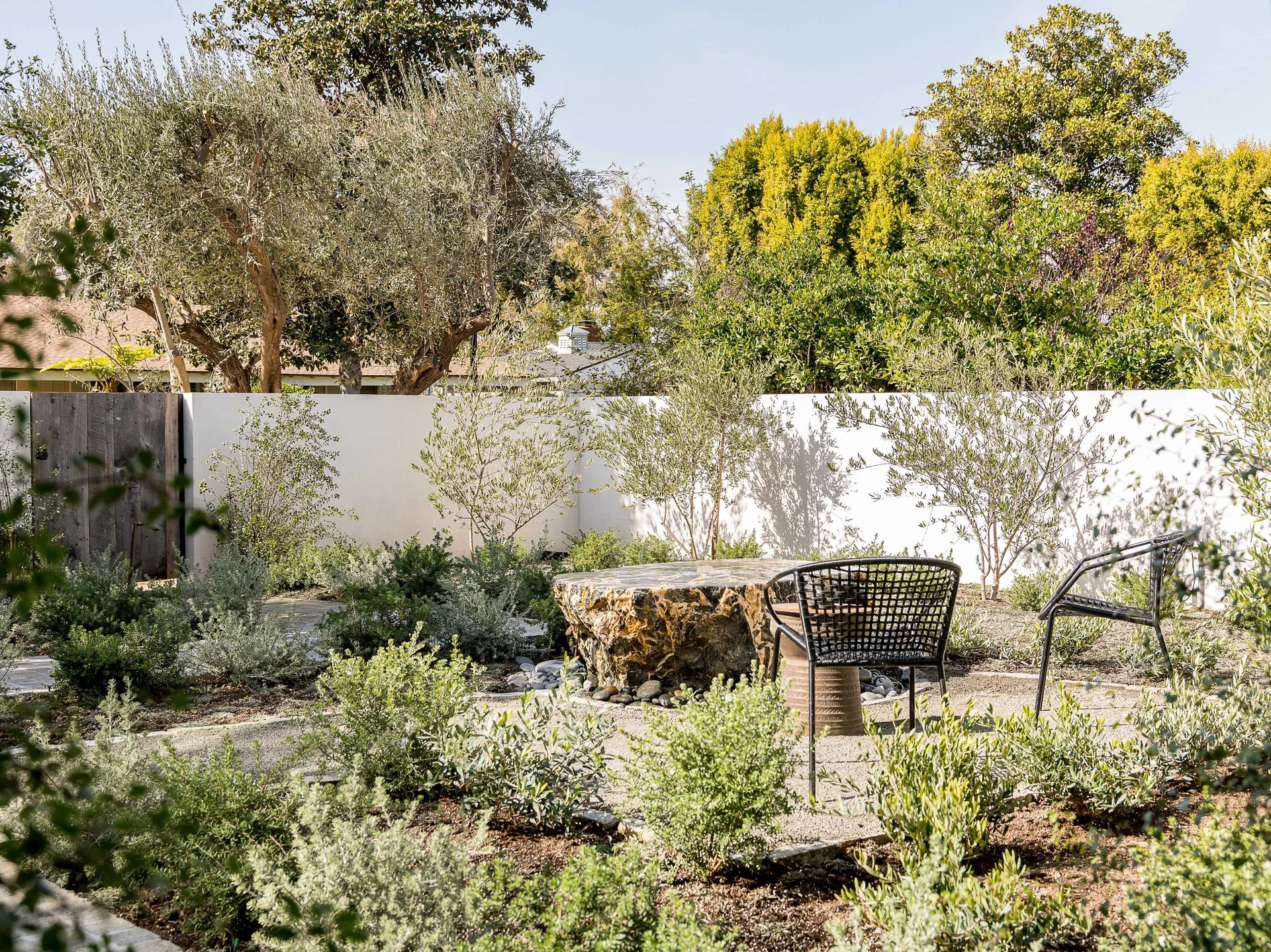 A landscaped garden features a large natural stone boulder table and black metal chairs set on a gravel path against a white boundary wall.