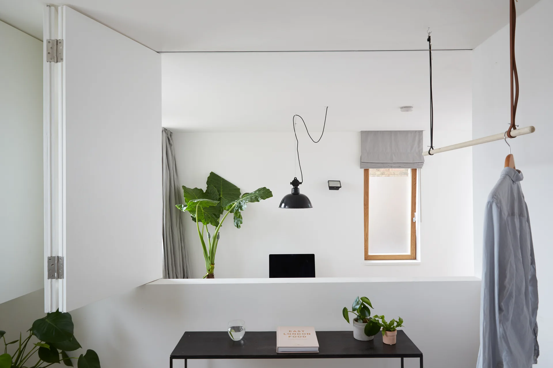 A minimalist white room featuring a low half-wall divider, a black console table with plants, and a wooden hanging clothes rack.