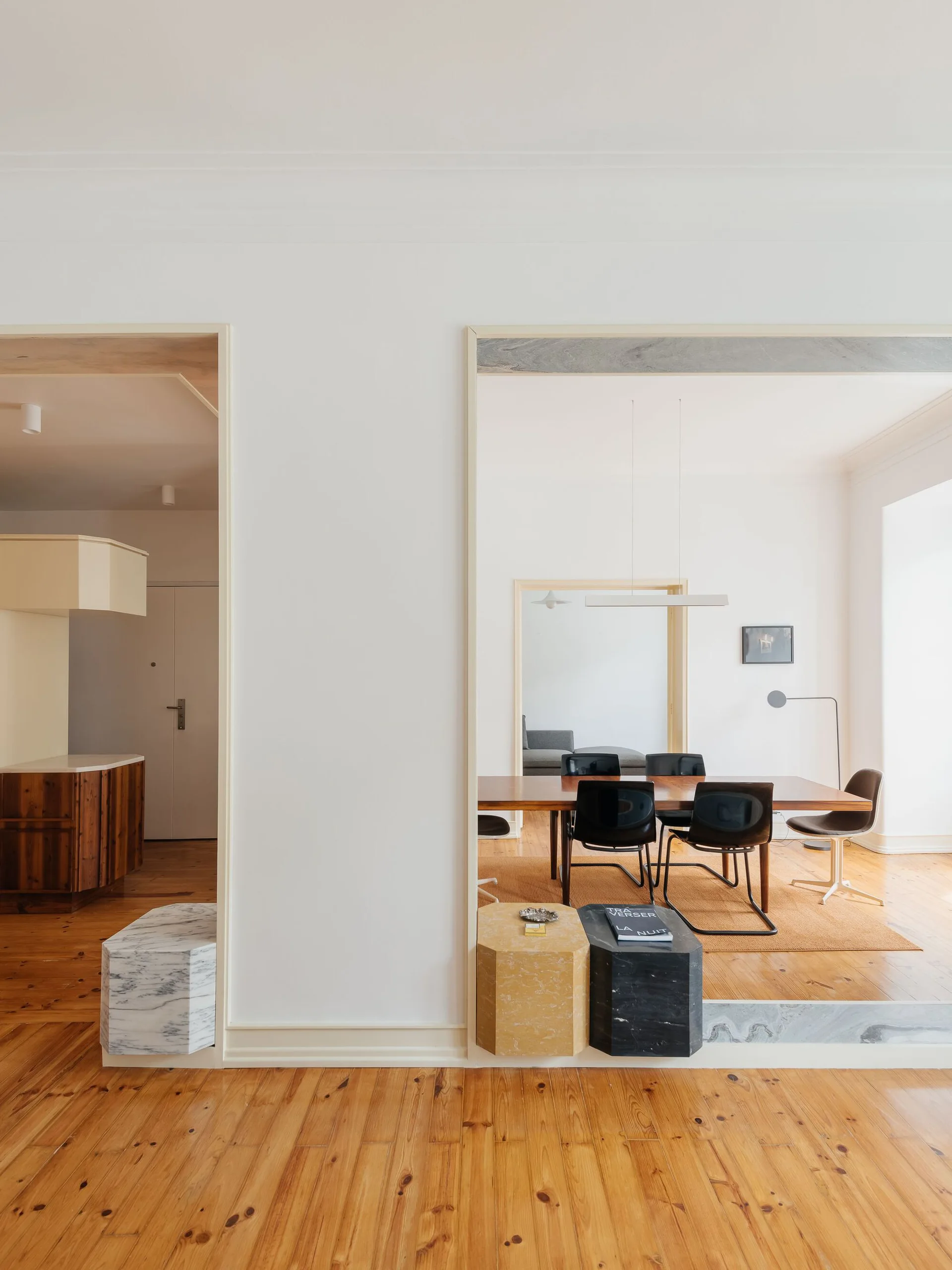 View through white wall openings framed with stone plinths into a dining room with a wooden table, black chairs, and wood flooring.