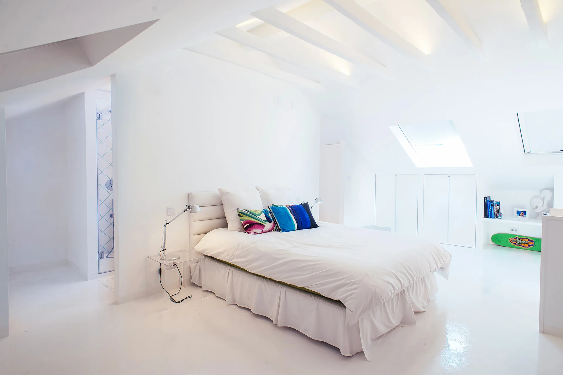 A minimalist white attic bedroom features a bed with colorful throw pillows, exposed ceiling beams, a skylight, and a clear acrylic bedside table.