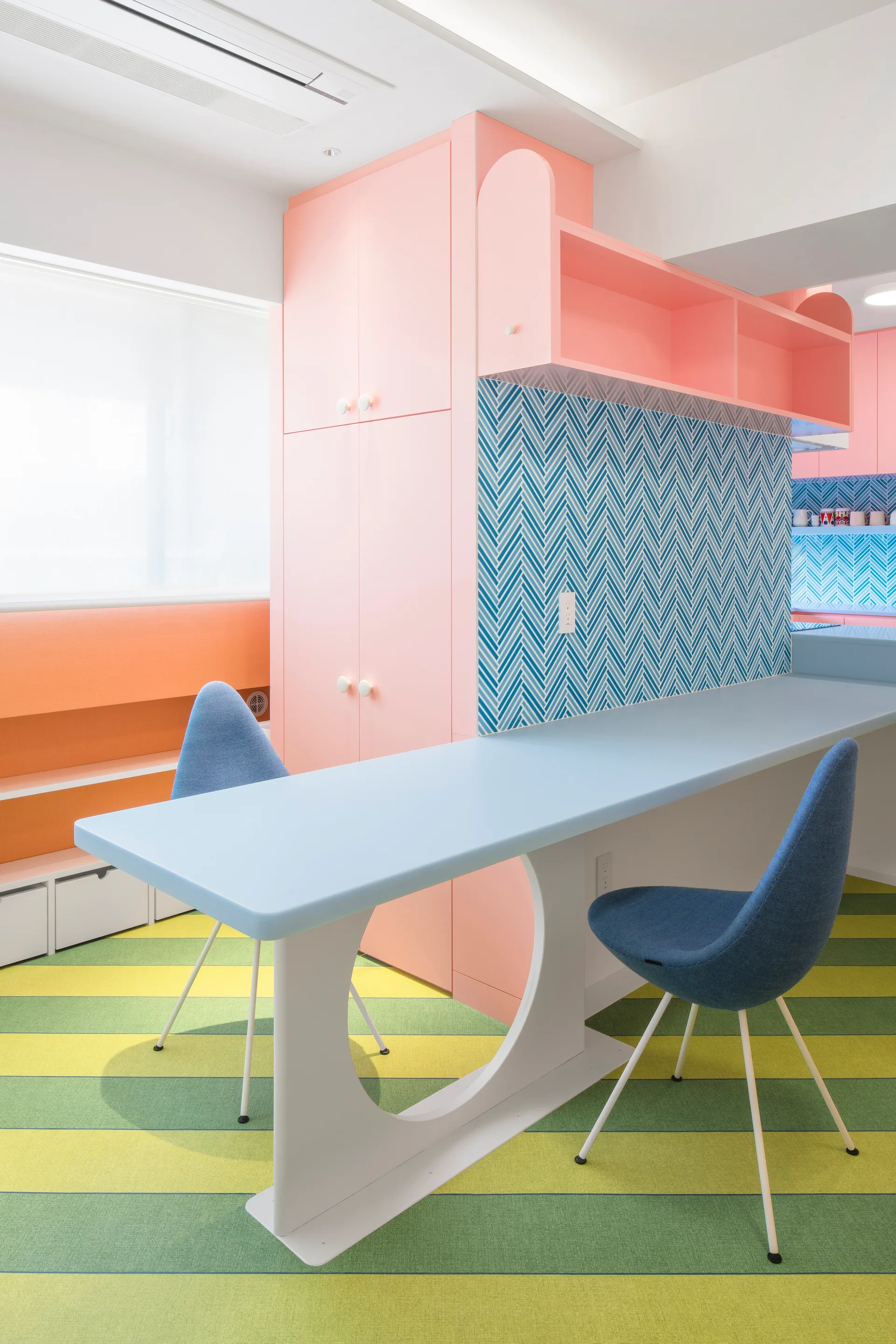 A modern kitchen features pastel pink cabinets, a light blue breakfast bar with blue upholstered chairs, and green striped flooring.