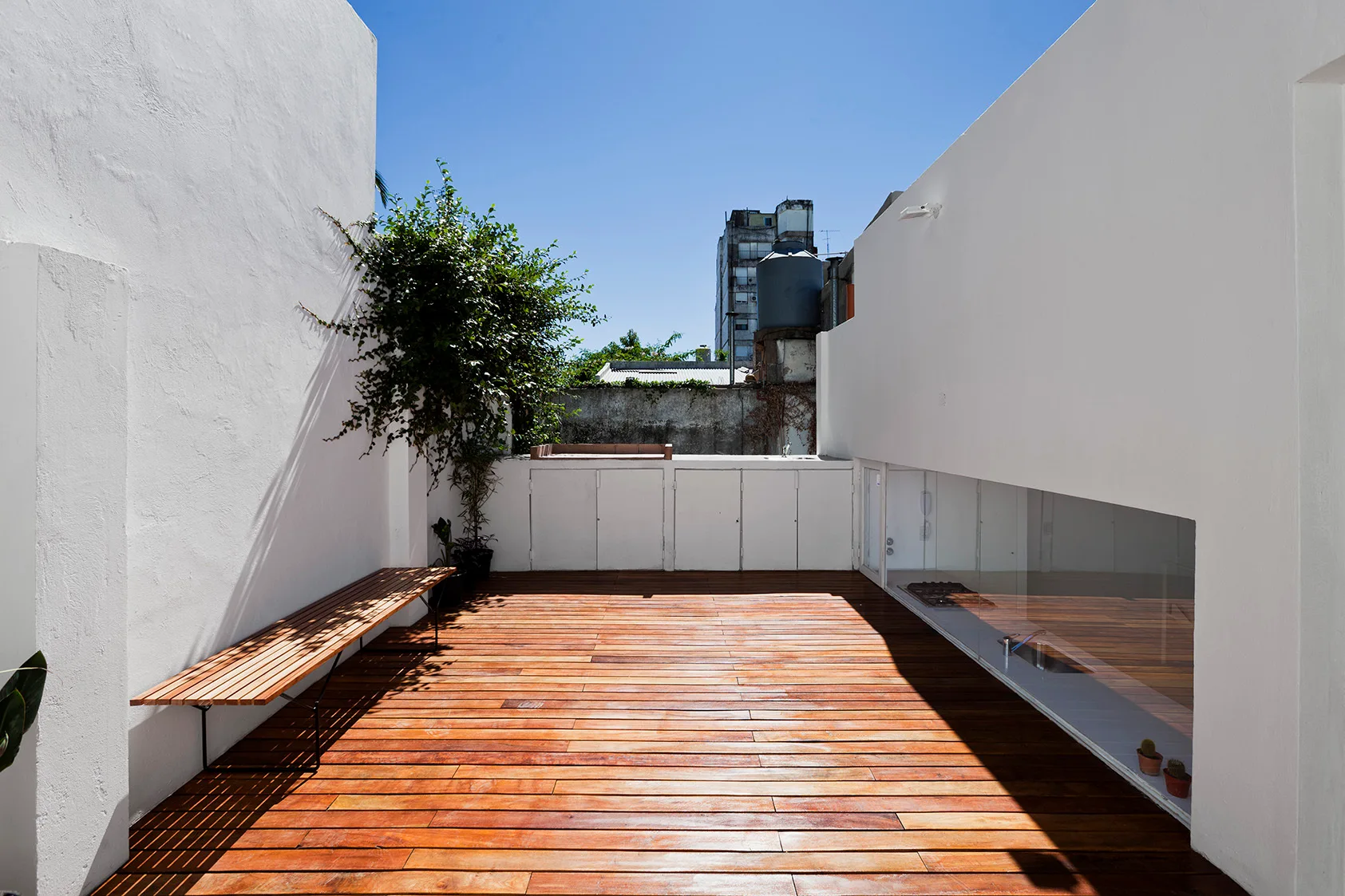 A minimalist outdoor terrace features warm wood plank decking, tall white walls, a wooden slatted bench, and a low glass window.