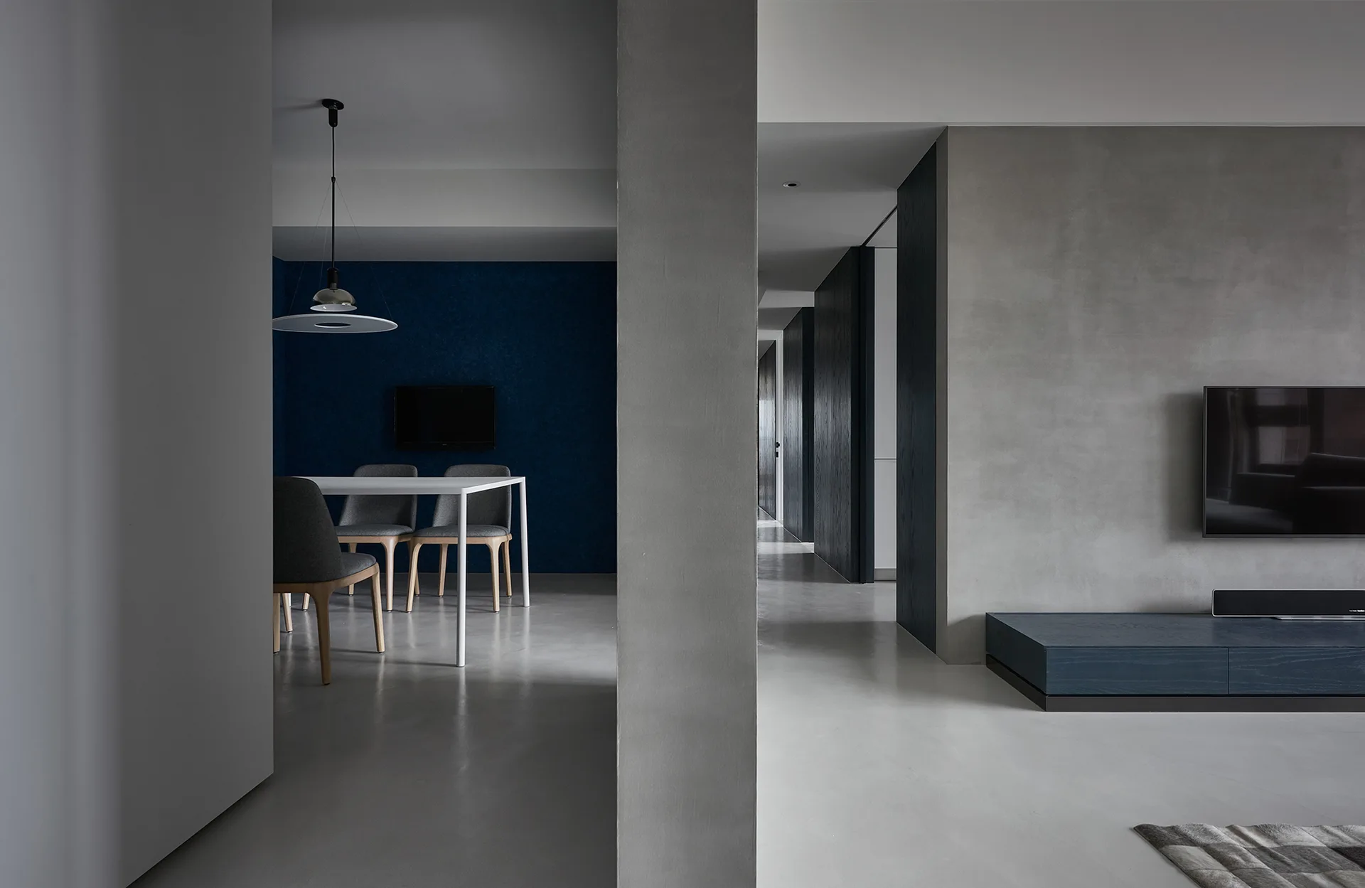 A minimalist open-plan living space features a gray concrete wall with a wall-mounted TV, adjacent to a dining area with a navy blue accent wall.
