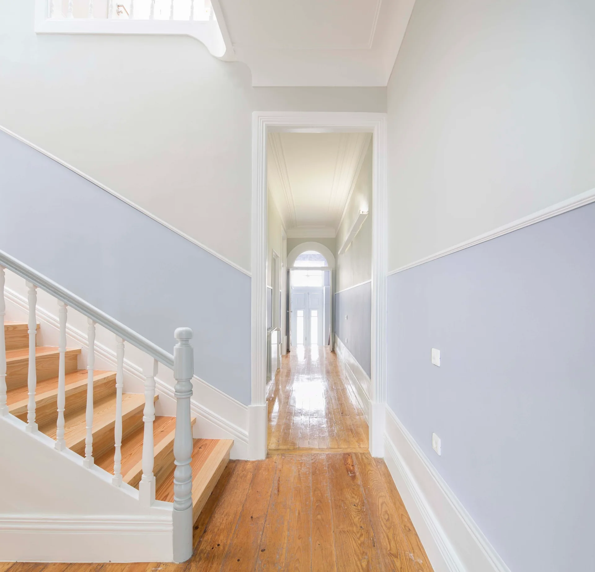 A hallway with blue and white two-toned walls, a wooden staircase, and polished pine wood flooring leading to an arched doorway.