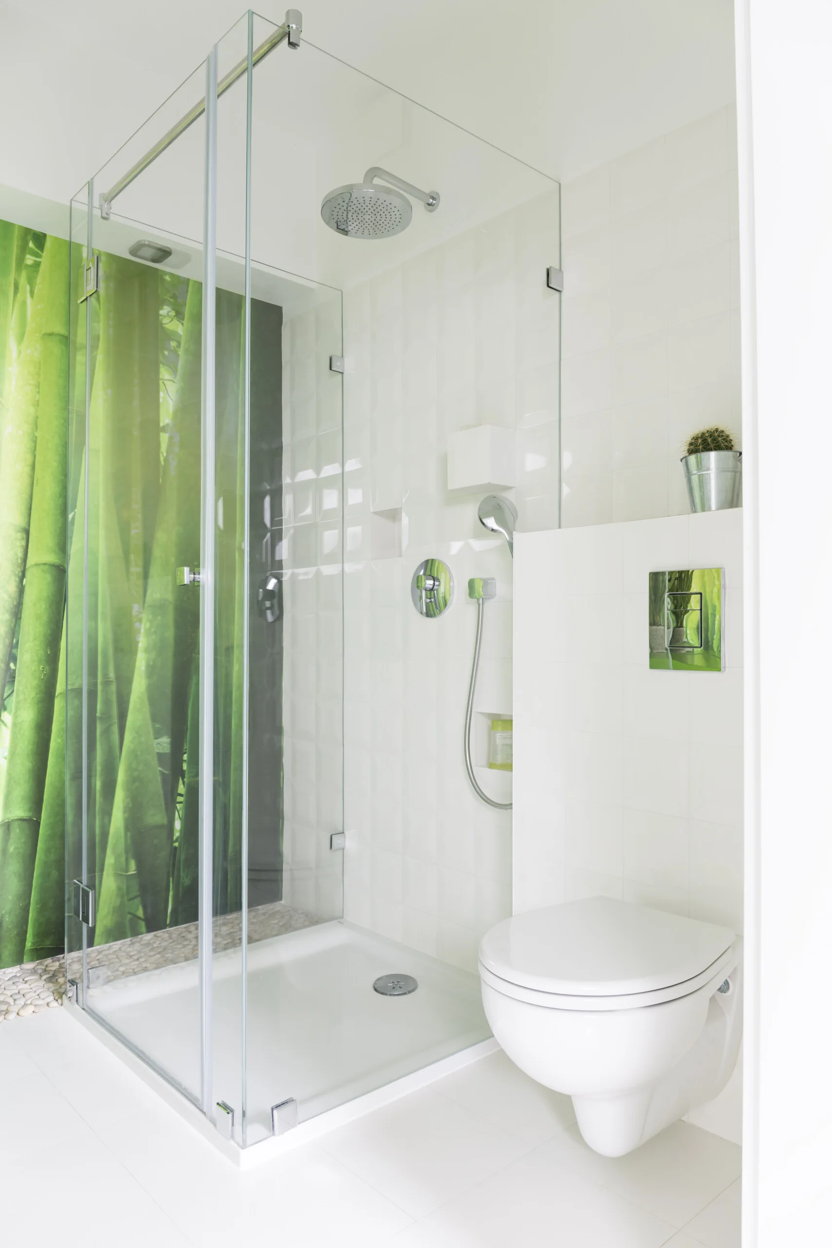 A modern white tiled bathroom featuring a glass-enclosed shower with a green bamboo print wall panel and a wall-mounted toilet.