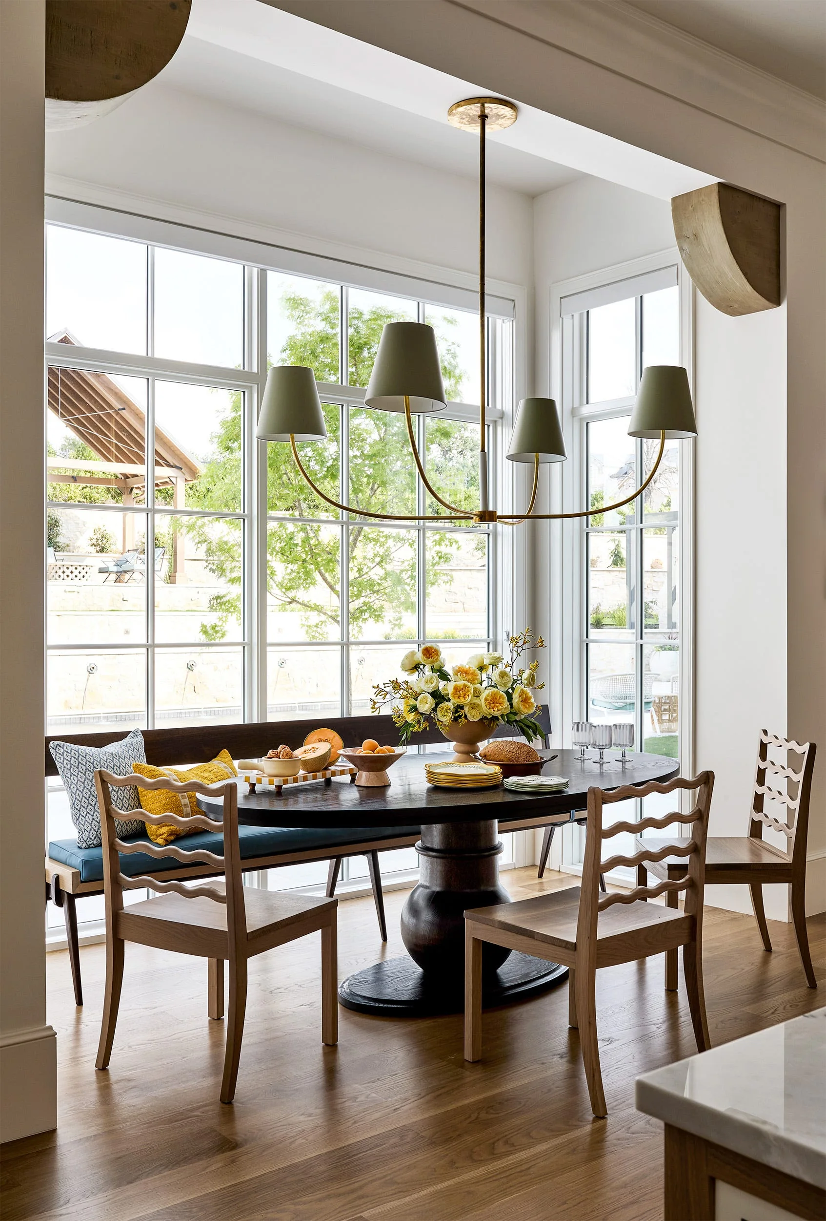 A dining area with a dark round wood table, curved wooden chairs, and a long blue cushioned banquette under a brass chandelier.