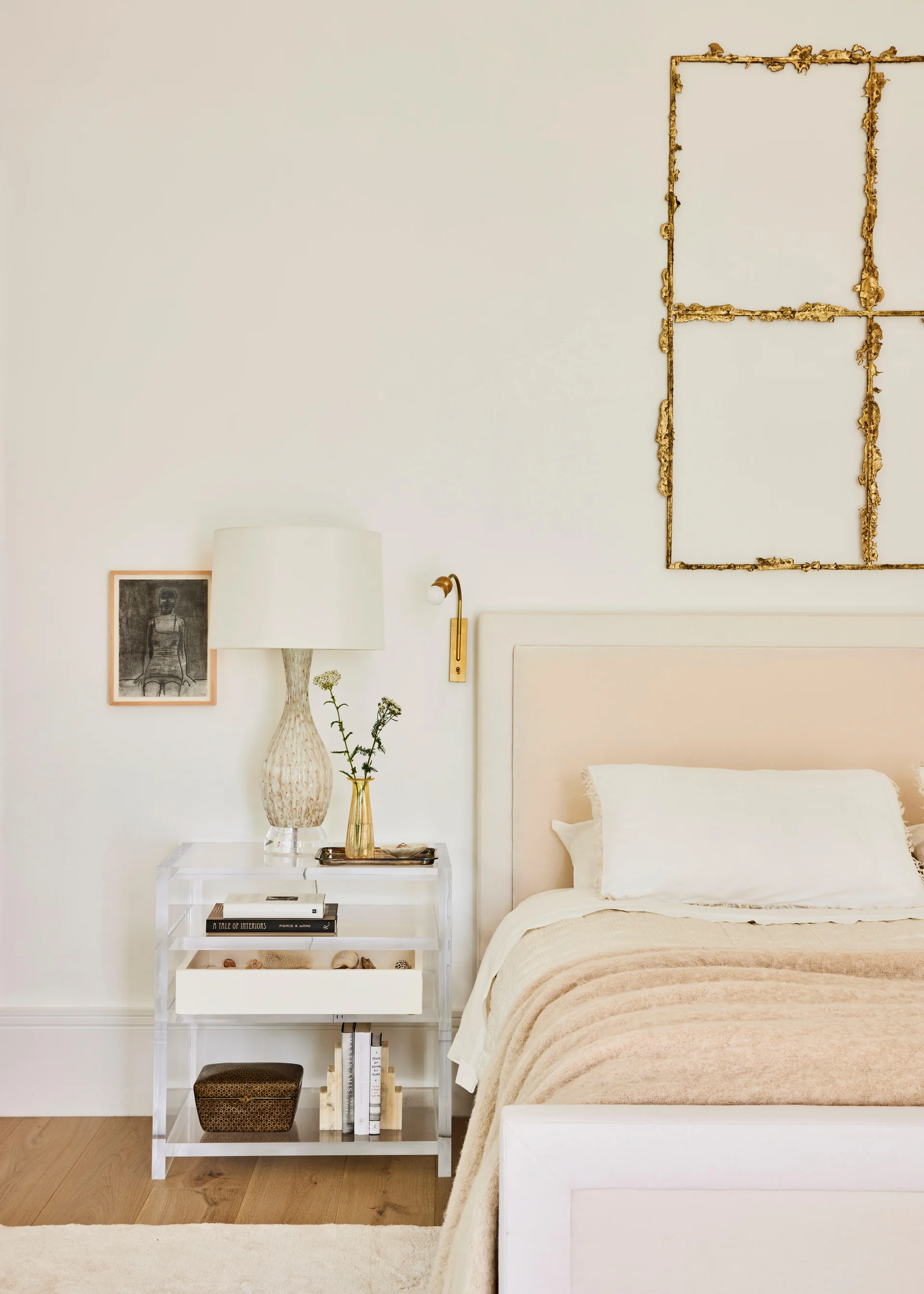 A neutral bedroom features a cream upholstered headboard, an acrylic bedside table with a lamp, and a gold metal sculpture on the wall.