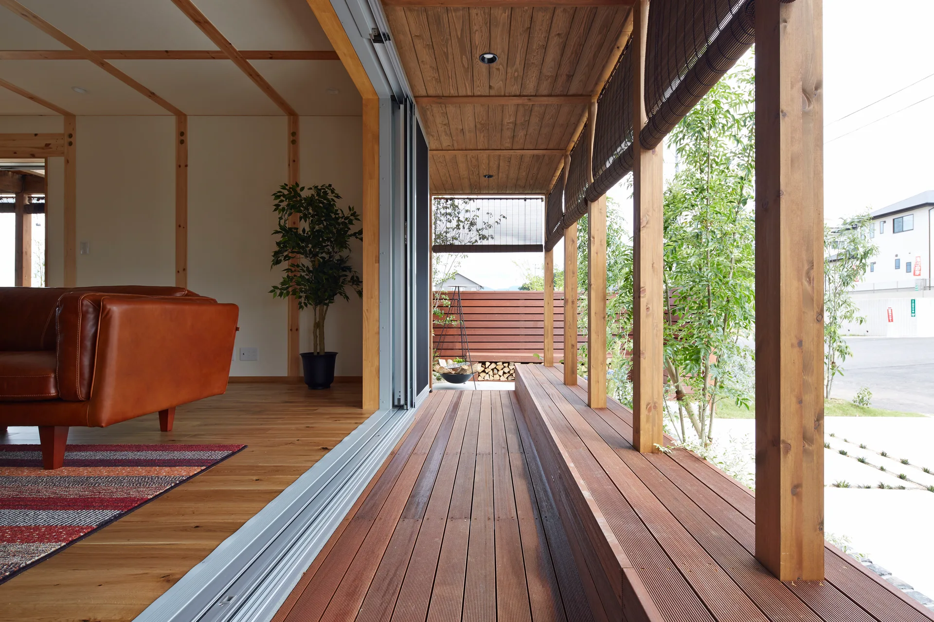 A large sliding glass door opens from a living room with a leather sofa onto a covered wooden deck with bamboo blinds.