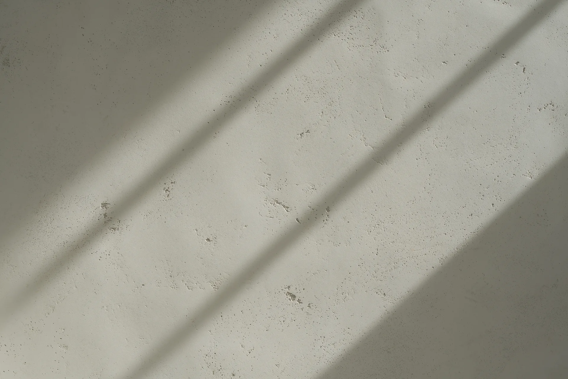 A close-up of a matte textured neutral plaster wall with small pitted details and diagonal shadows casting across the surface.