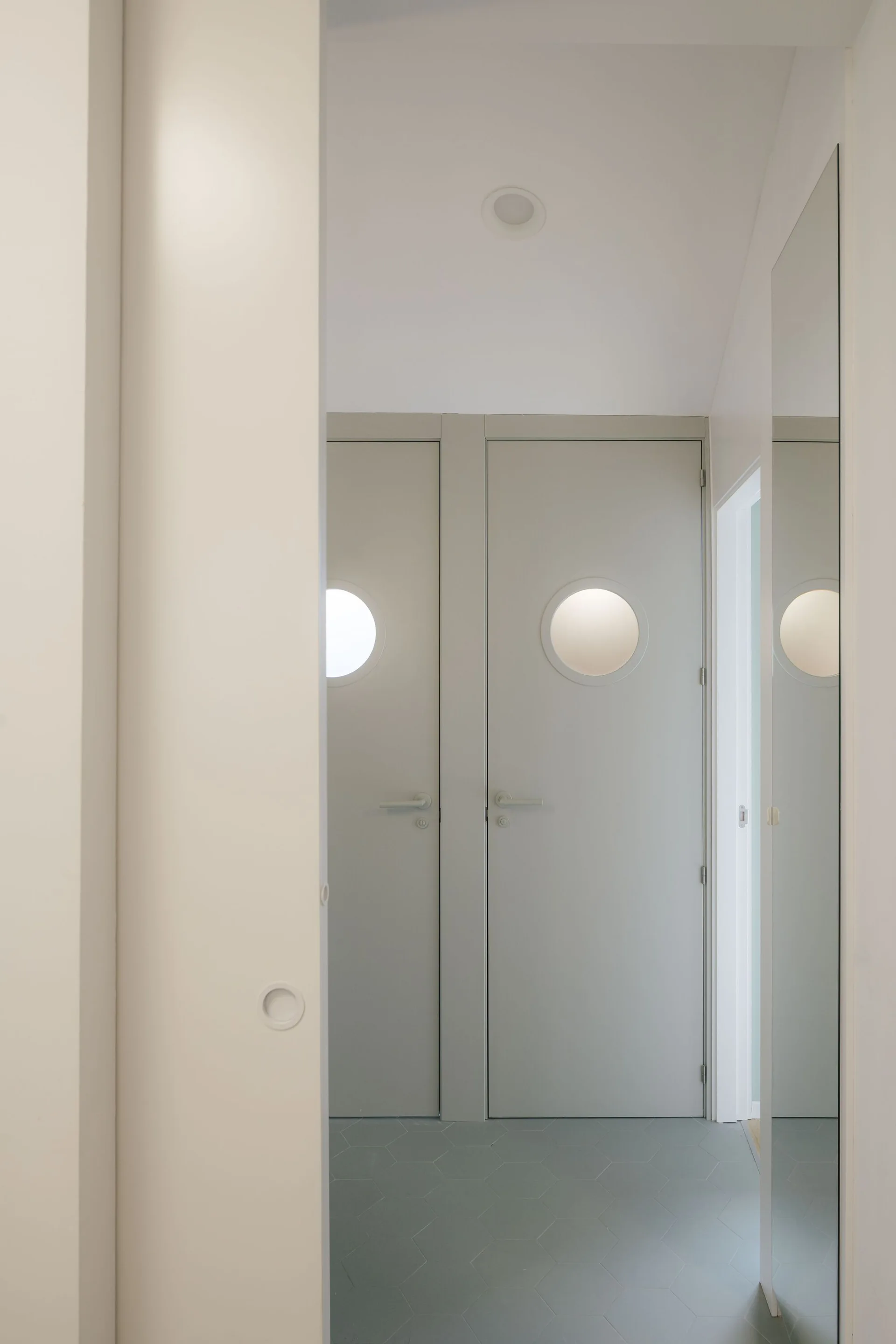 A minimalist hallway features two light grey doors with circular glass portholes and a grey hexagonal tile floor.