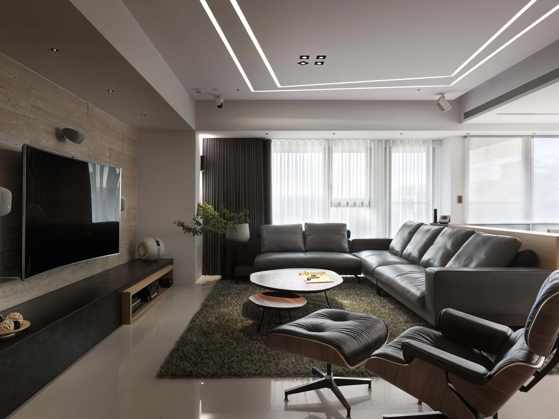 A modern living room features a grey leather sectional sofa, a classic lounge chair, nesting coffee tables, and recessed linear lighting in the ceiling.