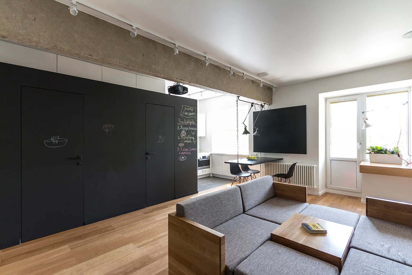 A modern living space featuring a grey sectional sofa, an exposed concrete ceiling beam, and a matte black chalkboard wall.