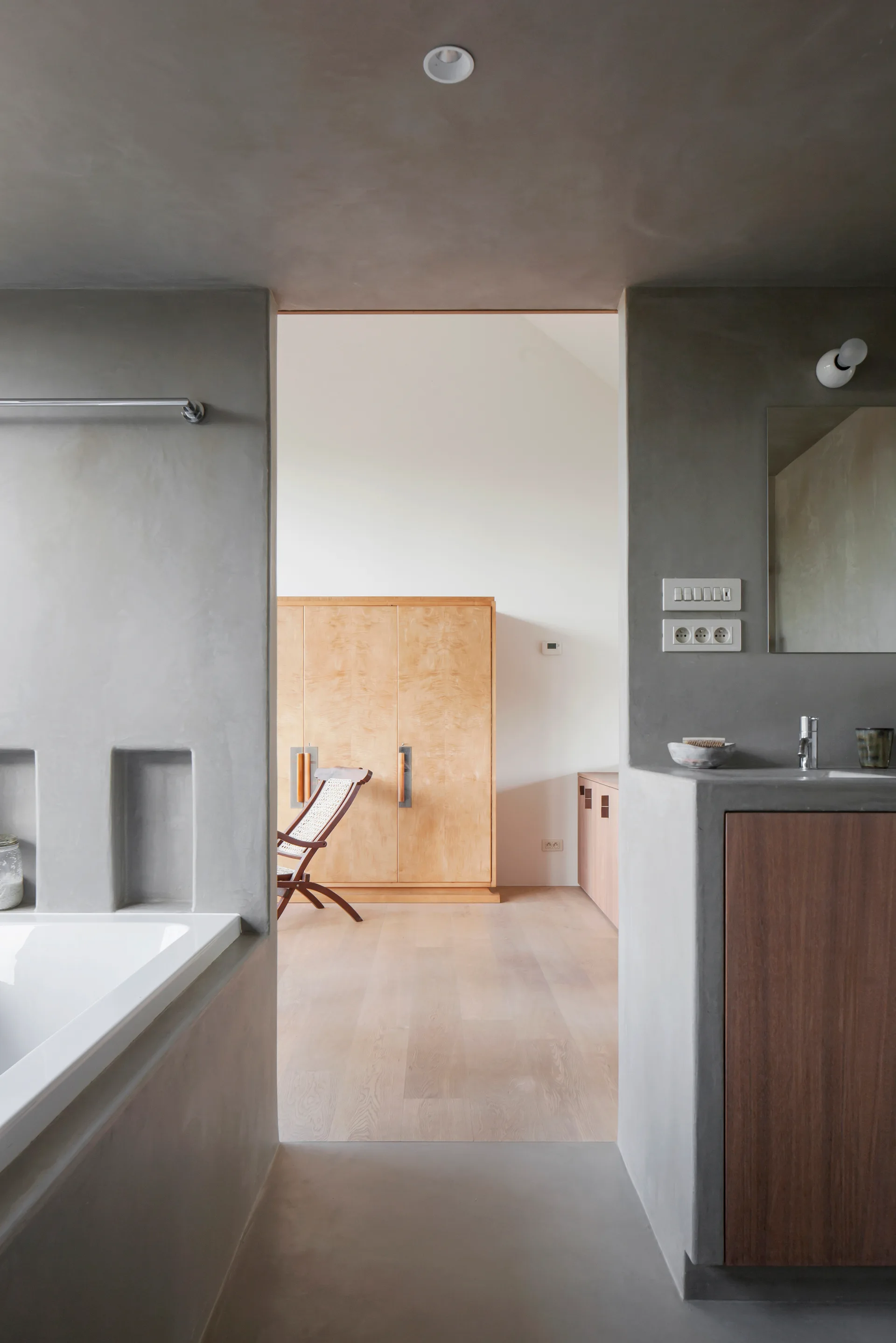 A grey plaster bathroom with an integrated concrete vanity and tub opens into a bright bedroom featuring a light wood wardrobe.