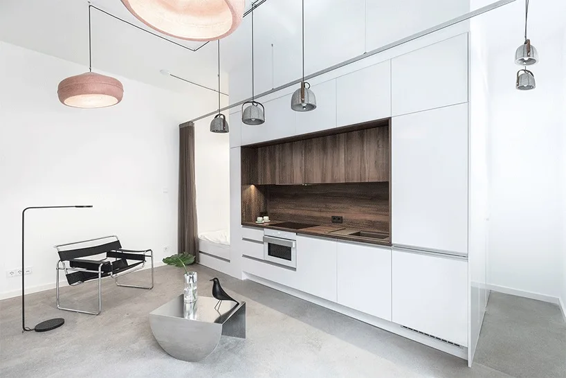 A minimalist studio kitchen featuring sleek white cabinetry with a warm recessed wood niche, a modern black armchair, and a chrome coffee table.