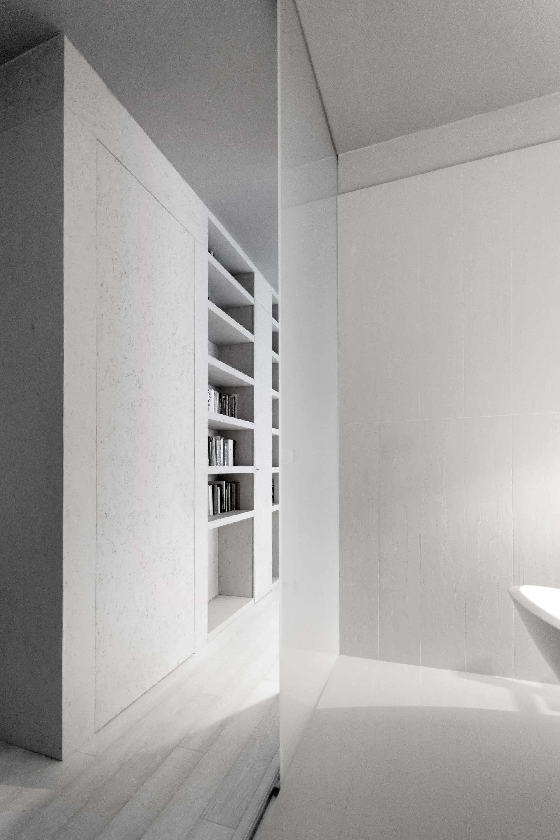 A minimalist white bathroom features a built-in floor-to-ceiling plaster bookshelf next to a glass partition and the edge of a white bathtub.