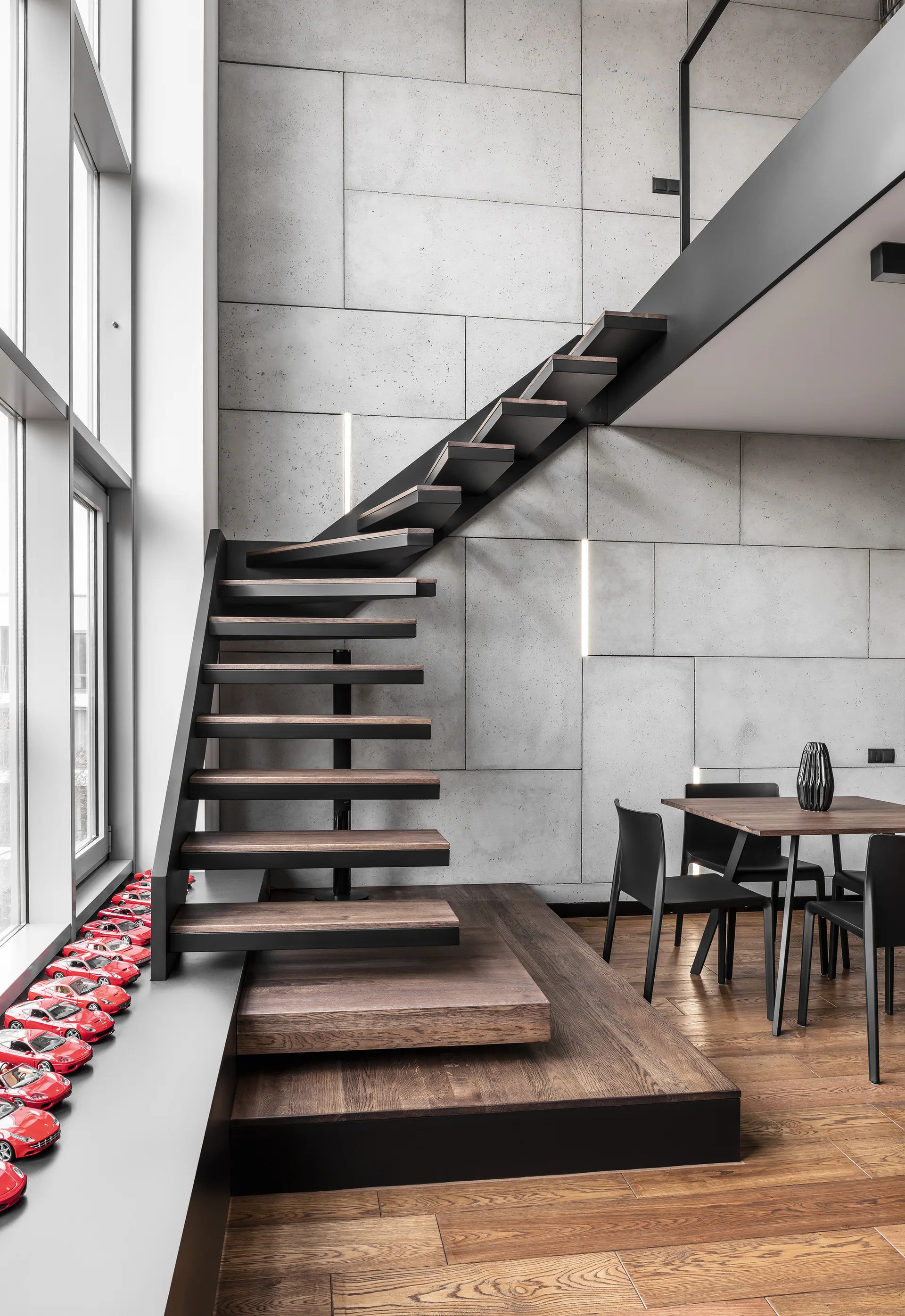 A modern floating staircase with dark wood treads sits against a gray concrete panel wall with vertical light strips next to a dining table.