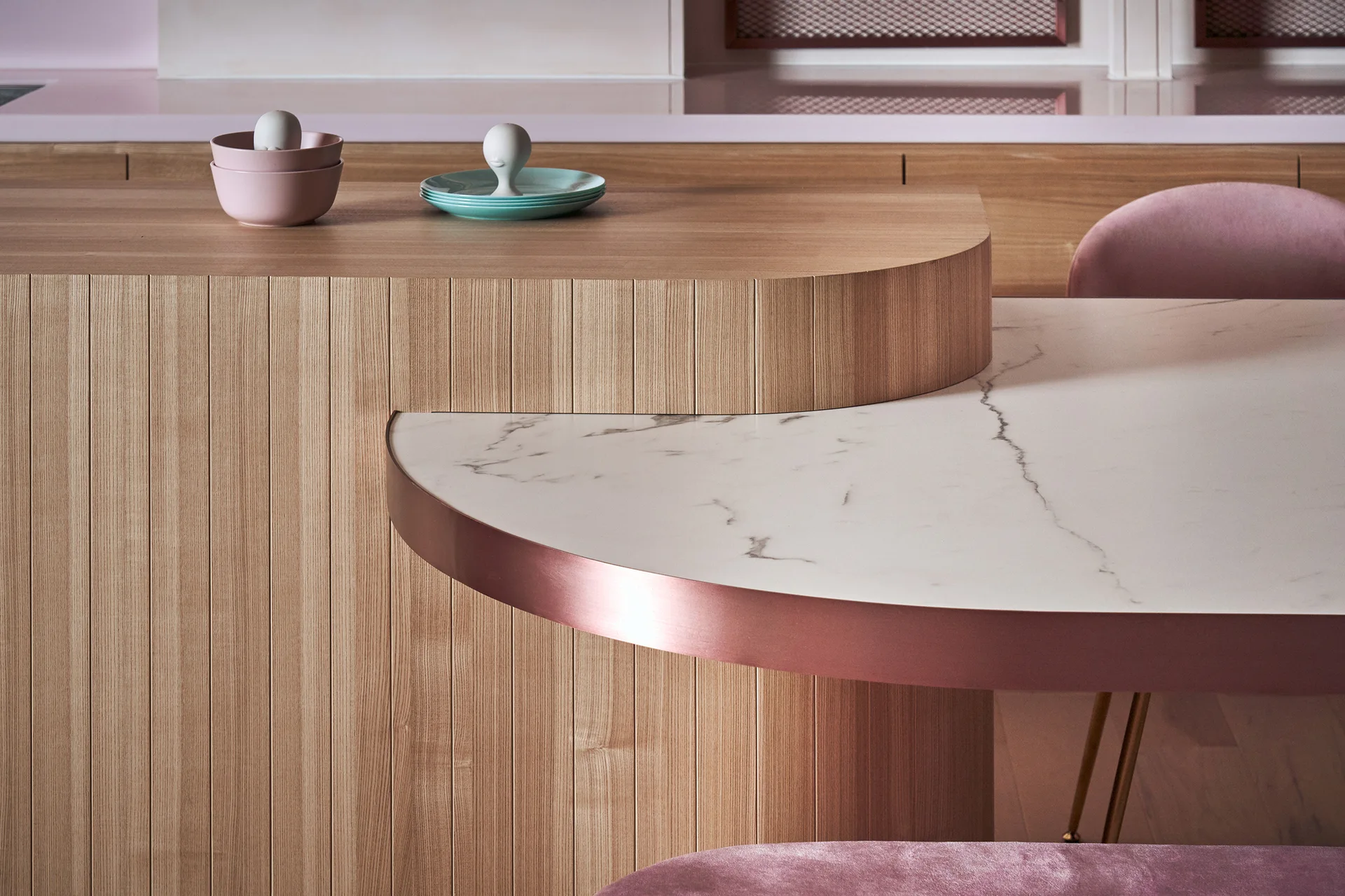 A close-up of a curved slatted wood kitchen island seamlessly connected to a round marble dining table with rose gold metallic trim and pink chairs.