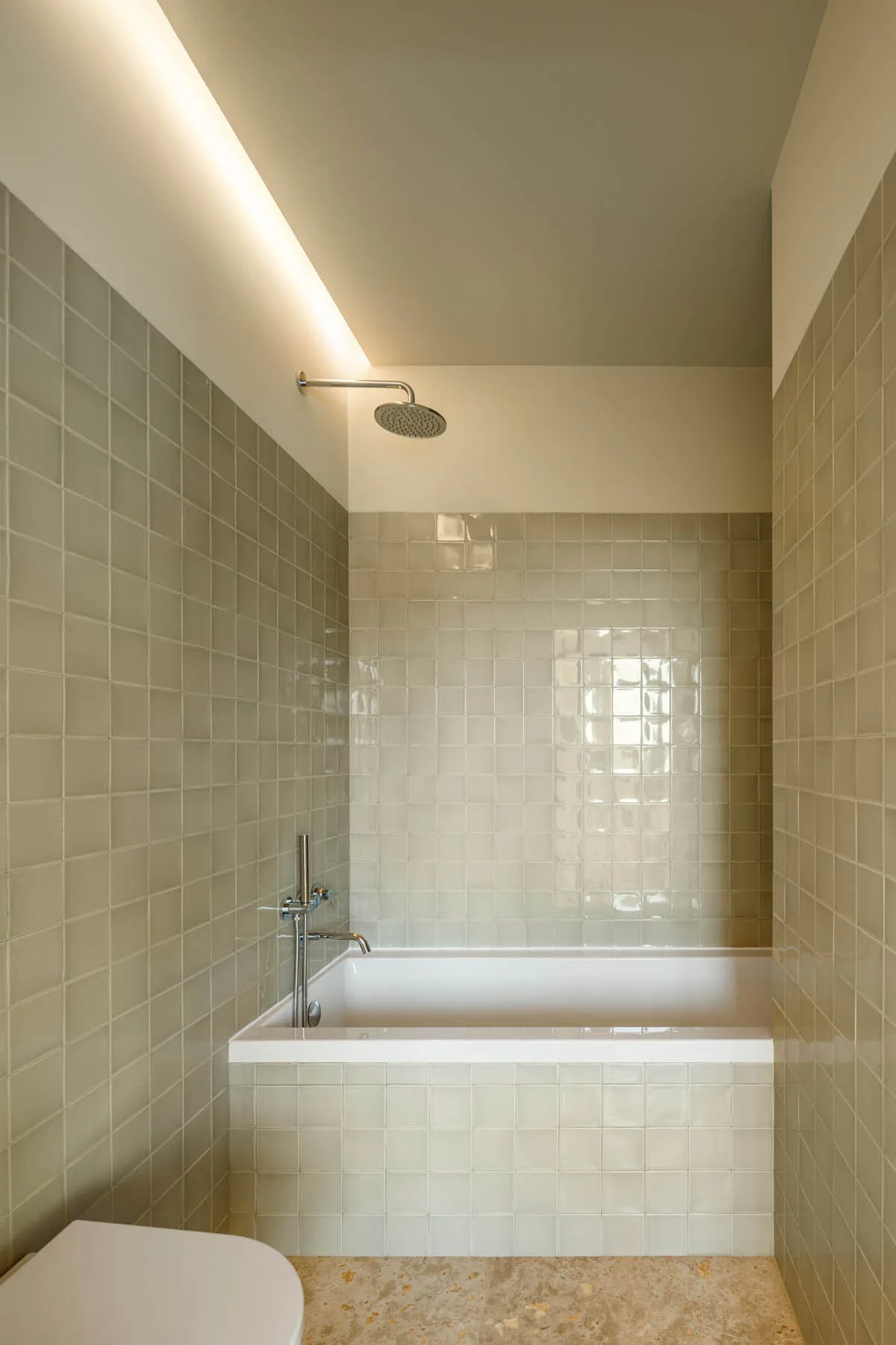 A minimalist bathroom alcove features pale green square glazed ceramic tiles, a white bathtub, chrome fixtures, and warm overhead cove lighting.