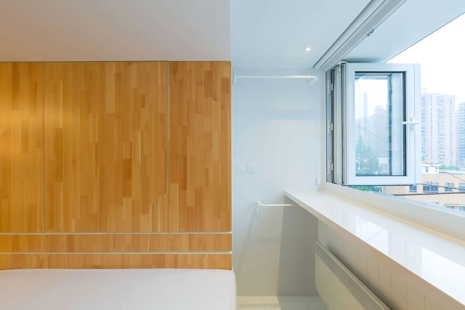 A minimalist bedroom featuring light wood wall paneling next to a bright white wall with an open window and a long built-in ledge.